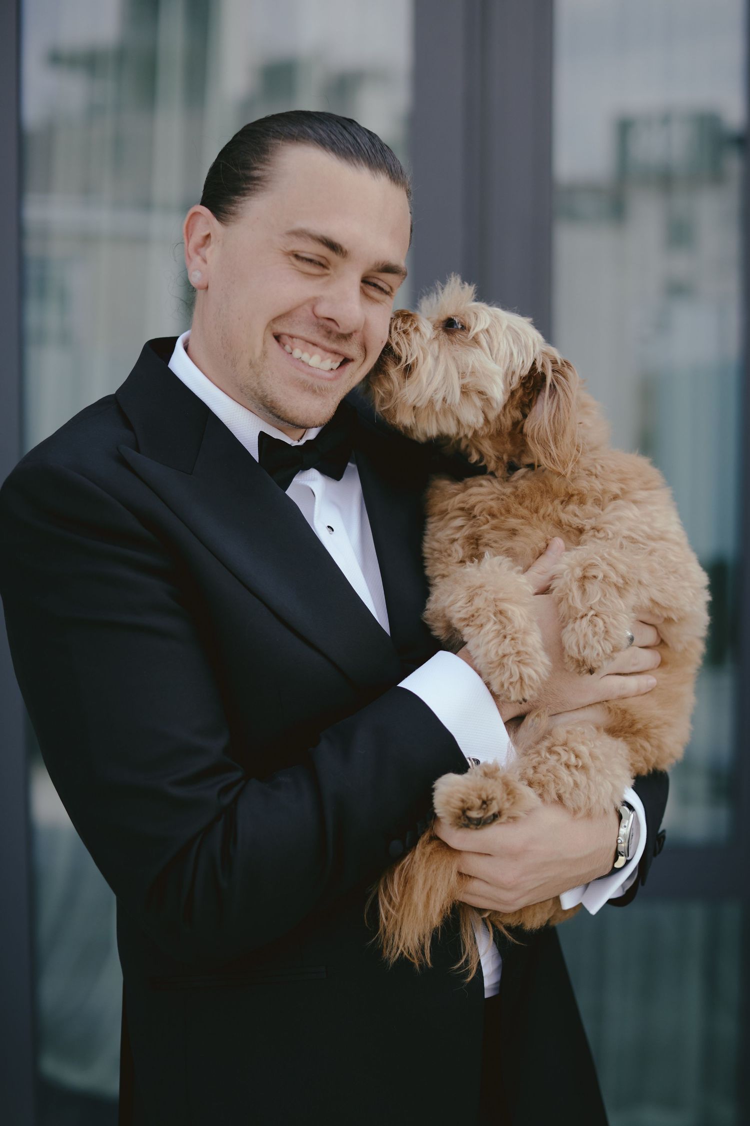 A person in a black tuxedo joyfully holds and embraces a fluffy golden-brown dog against a modern building backdrop.