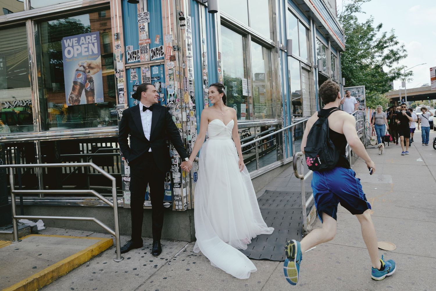 A couple in formal wedding attire stands outside a storefront as a jogger runs past on a city sidewalk.