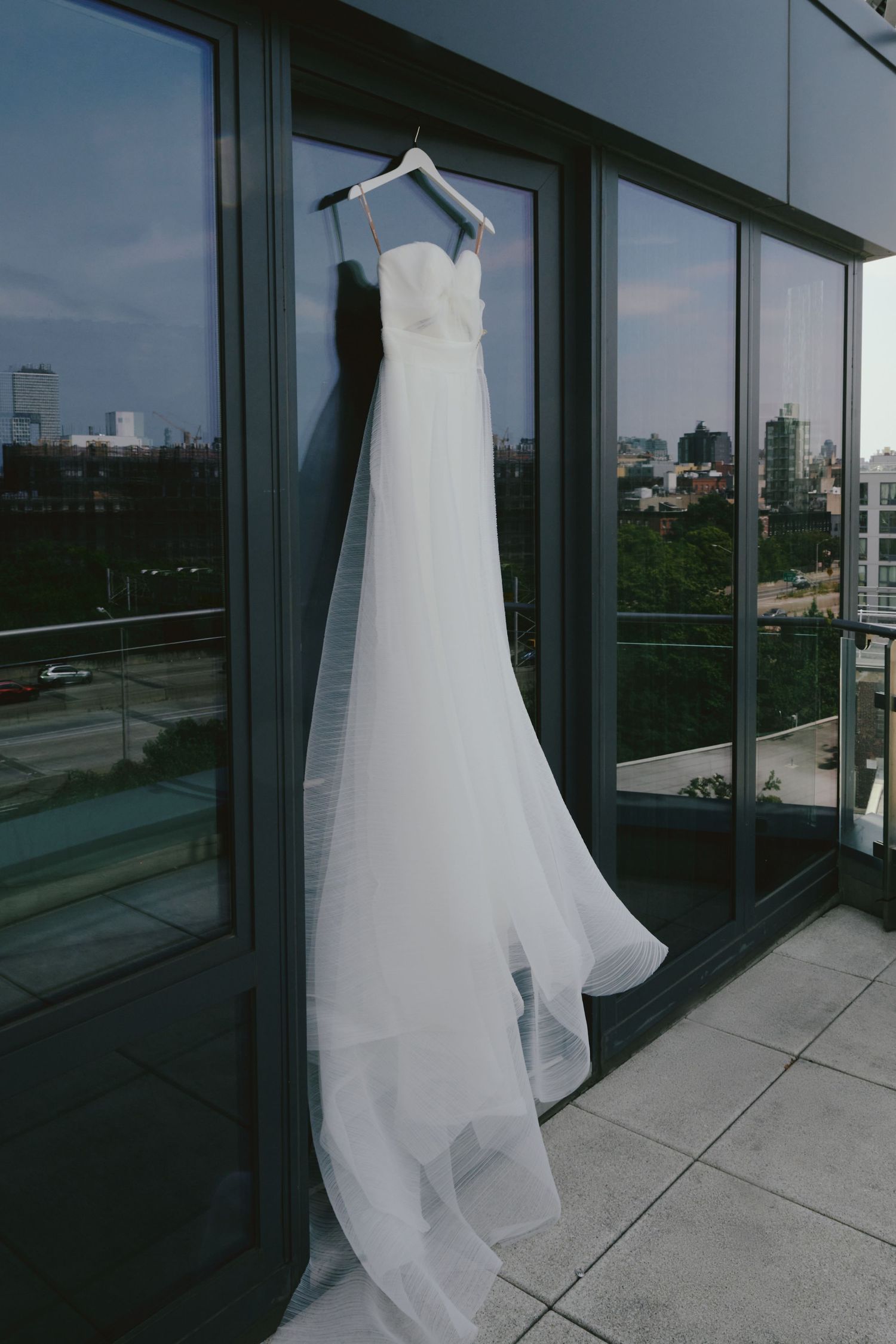 A white wedding dress hanging on a balcony with city views, showcasing its elegant flowing train and strapless design.