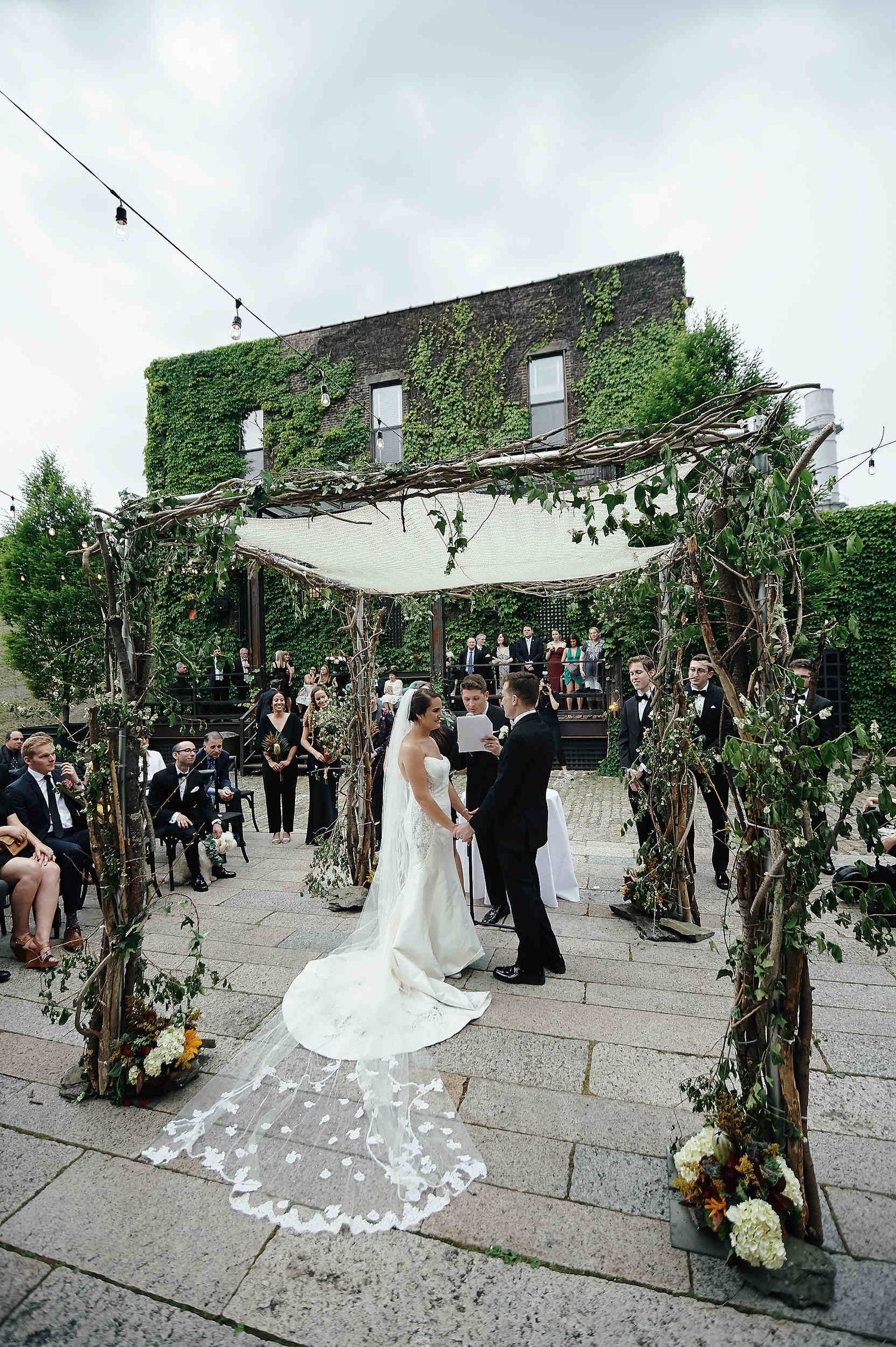 A wedding ceremony takes place at a rustic stone venue with ivy-covered walls and floral arches under a white canopy.