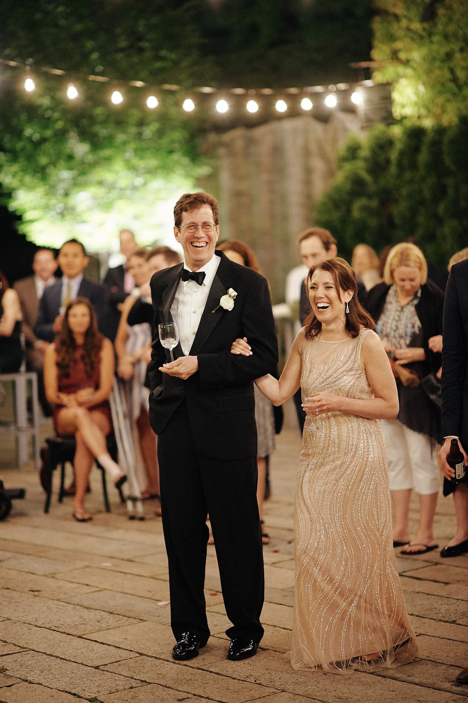 A joyous couple walking arm in arm at night under string lights at an outdoor wedding reception.