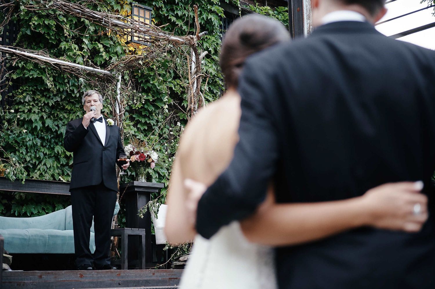A black-tie outdoor wedding ceremony with a couple and officiant surrounded by lush ivy-covered walls.