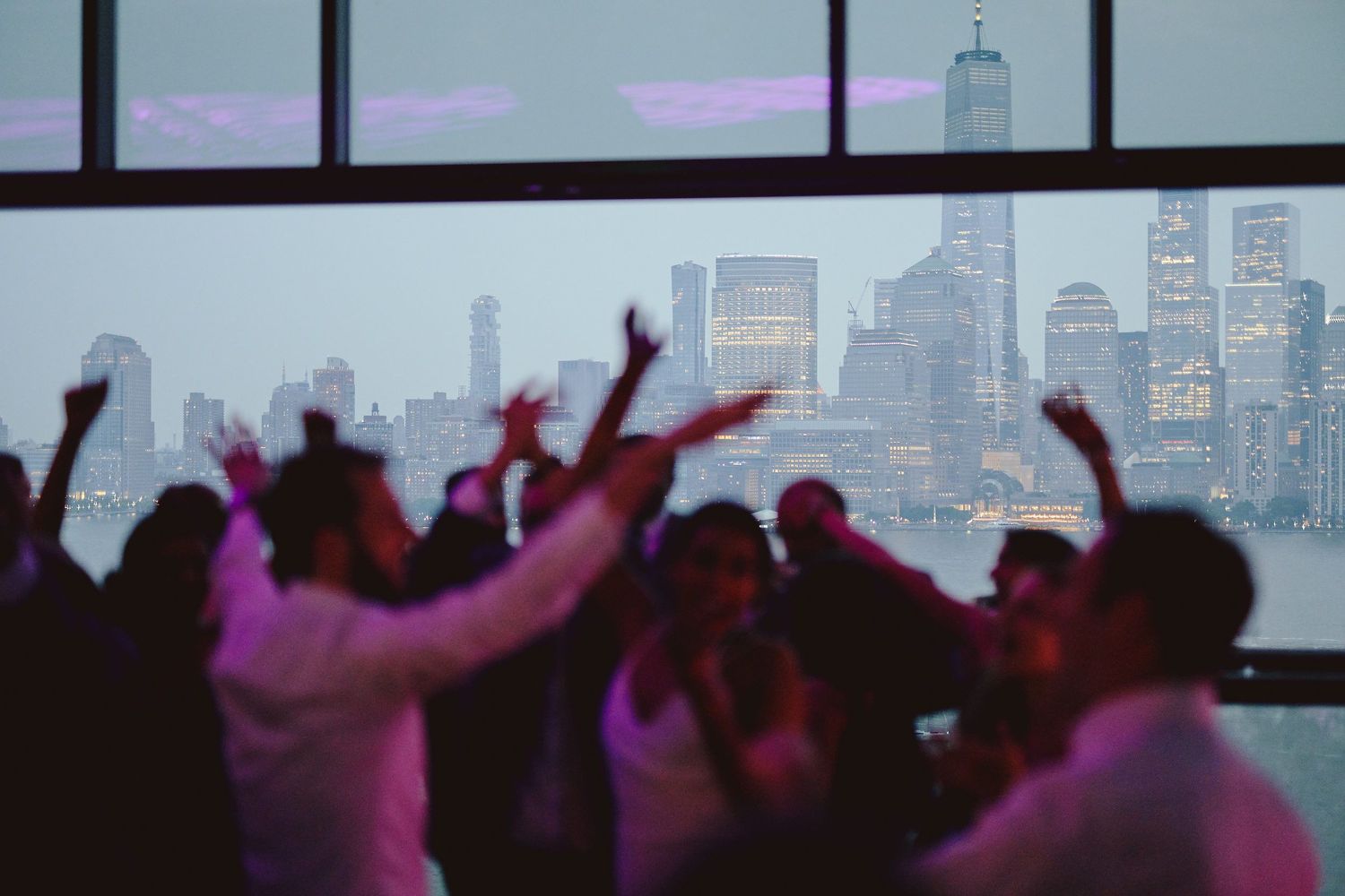 Silhouettes of party crowd dancing against New York City skyline with pink and purple lighting.