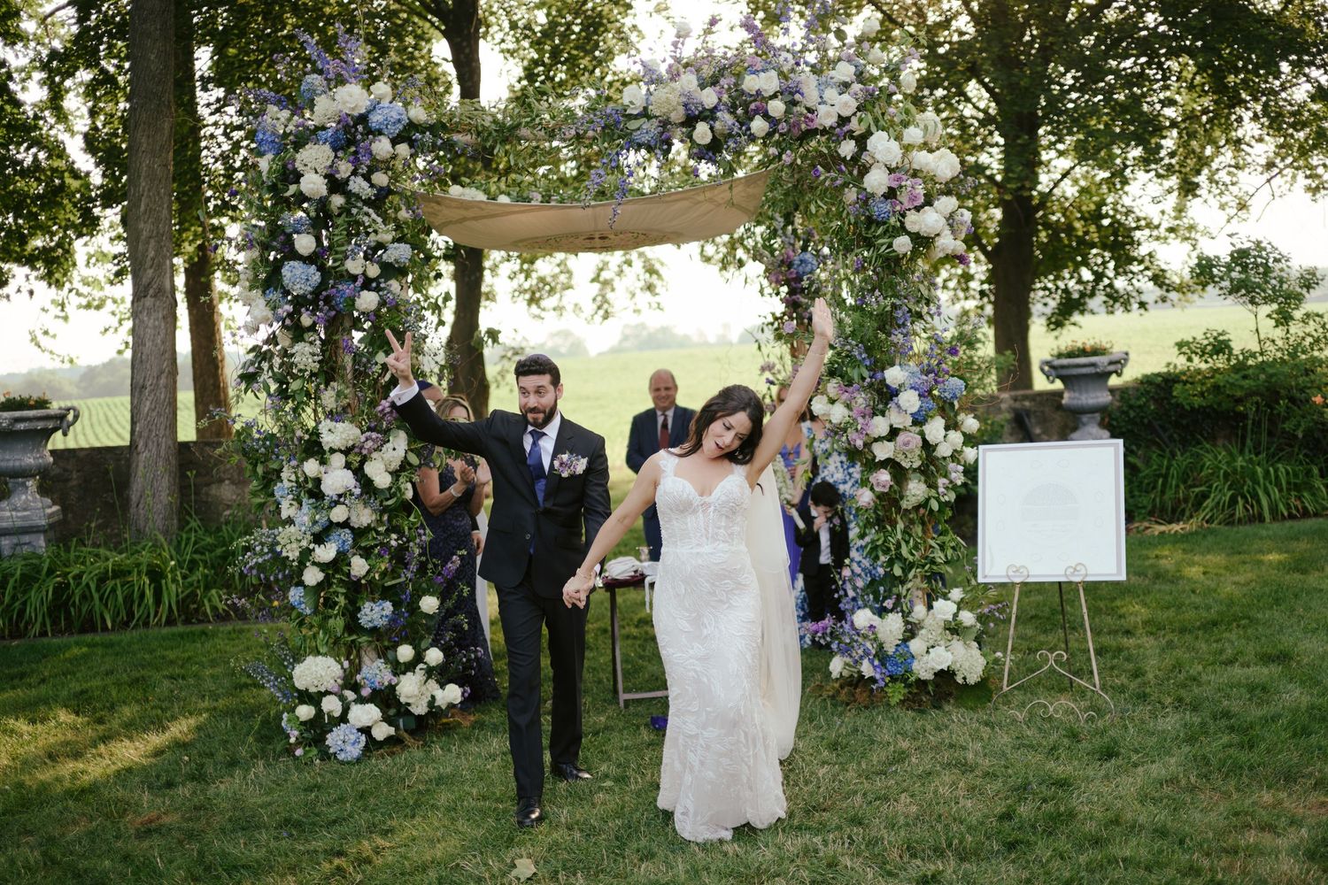 A couple celebrates their outdoor wedding ceremony with joyful gestures surrounded by floral arrangements on a grassy hill.