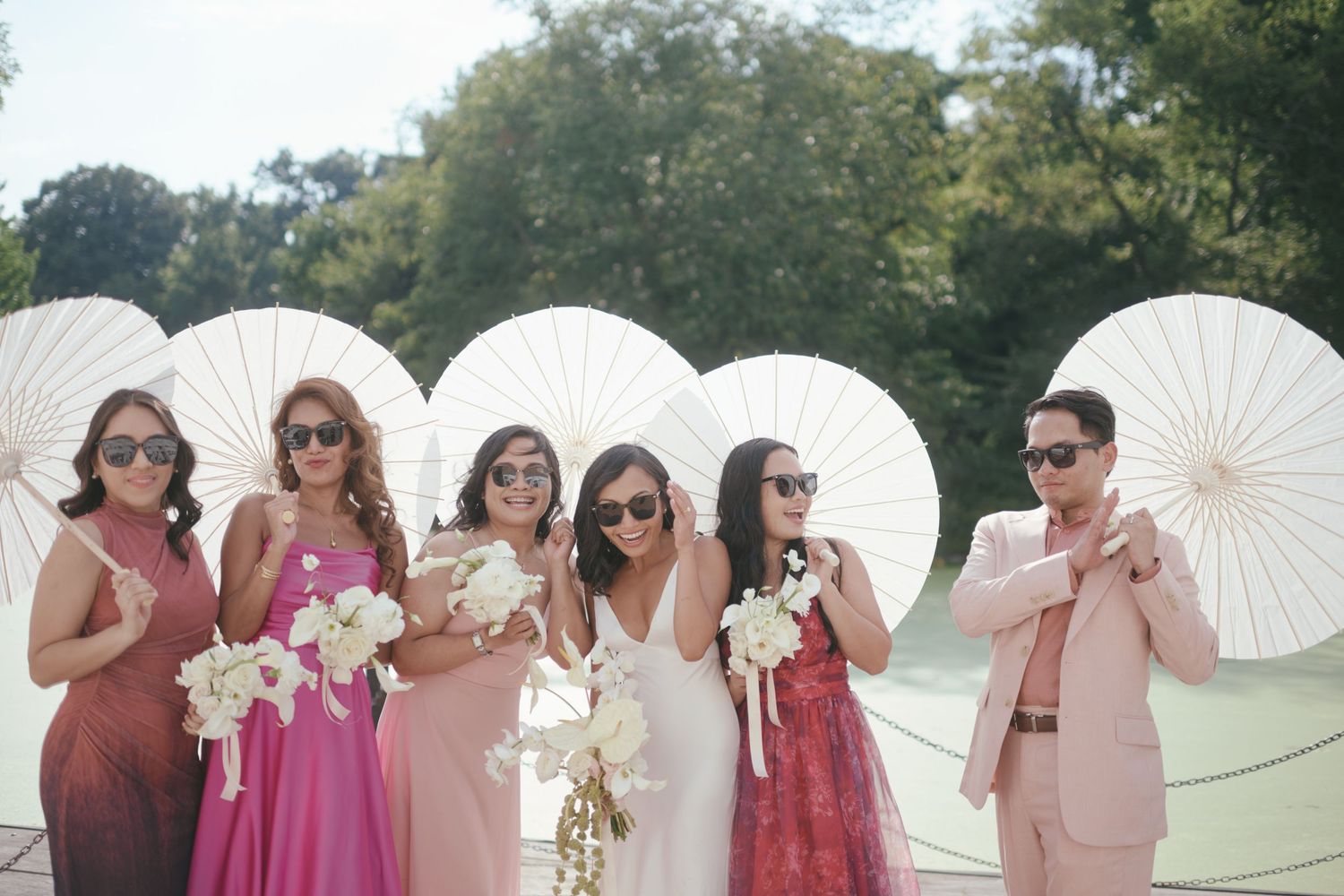 Bridal party in pink dresses holding white parasols and bouquets, posing outdoors with lush green trees behind them.