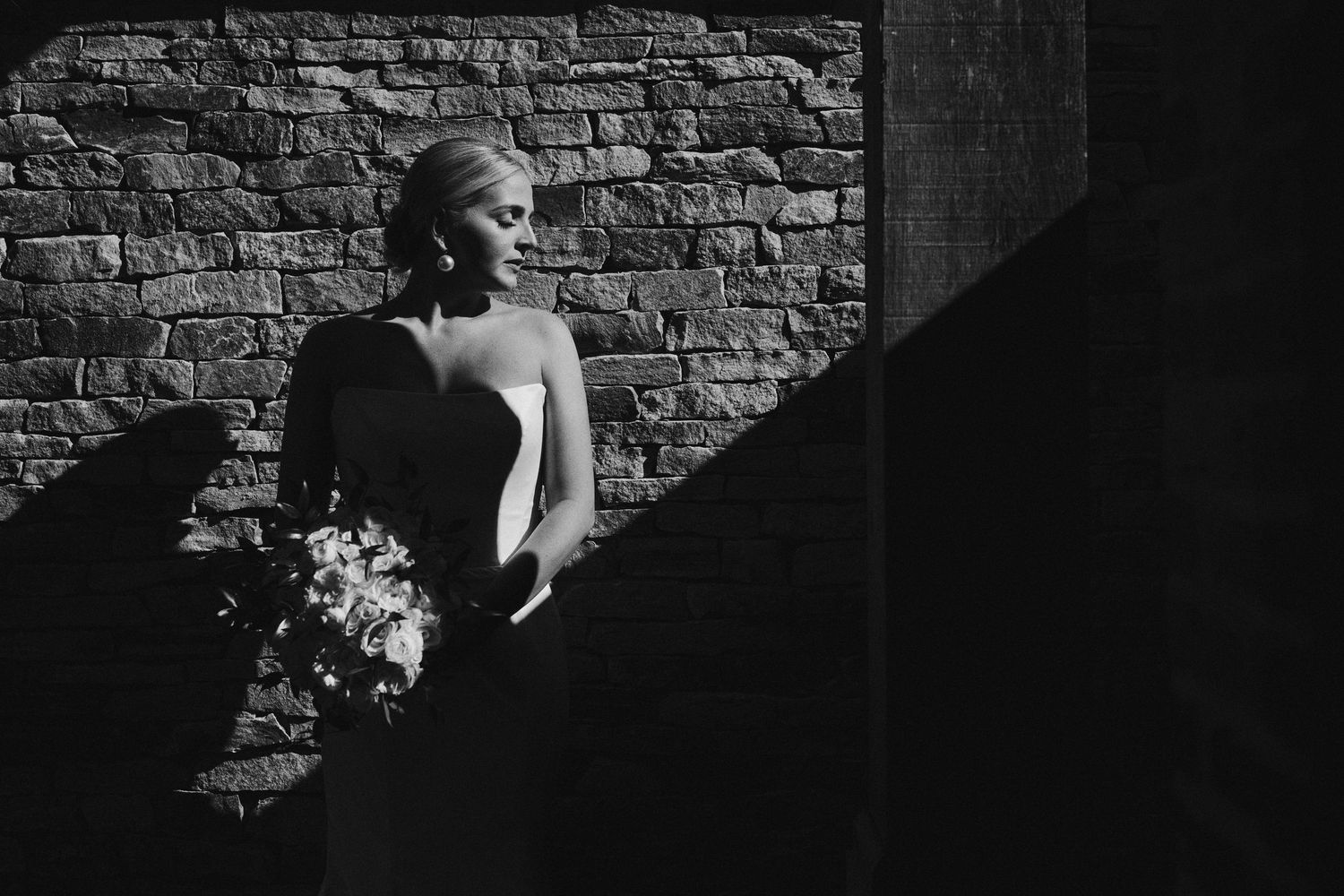 Black and white bridal portrait of elegant bride holding bouquet against rustic brick wall in dramatic light.