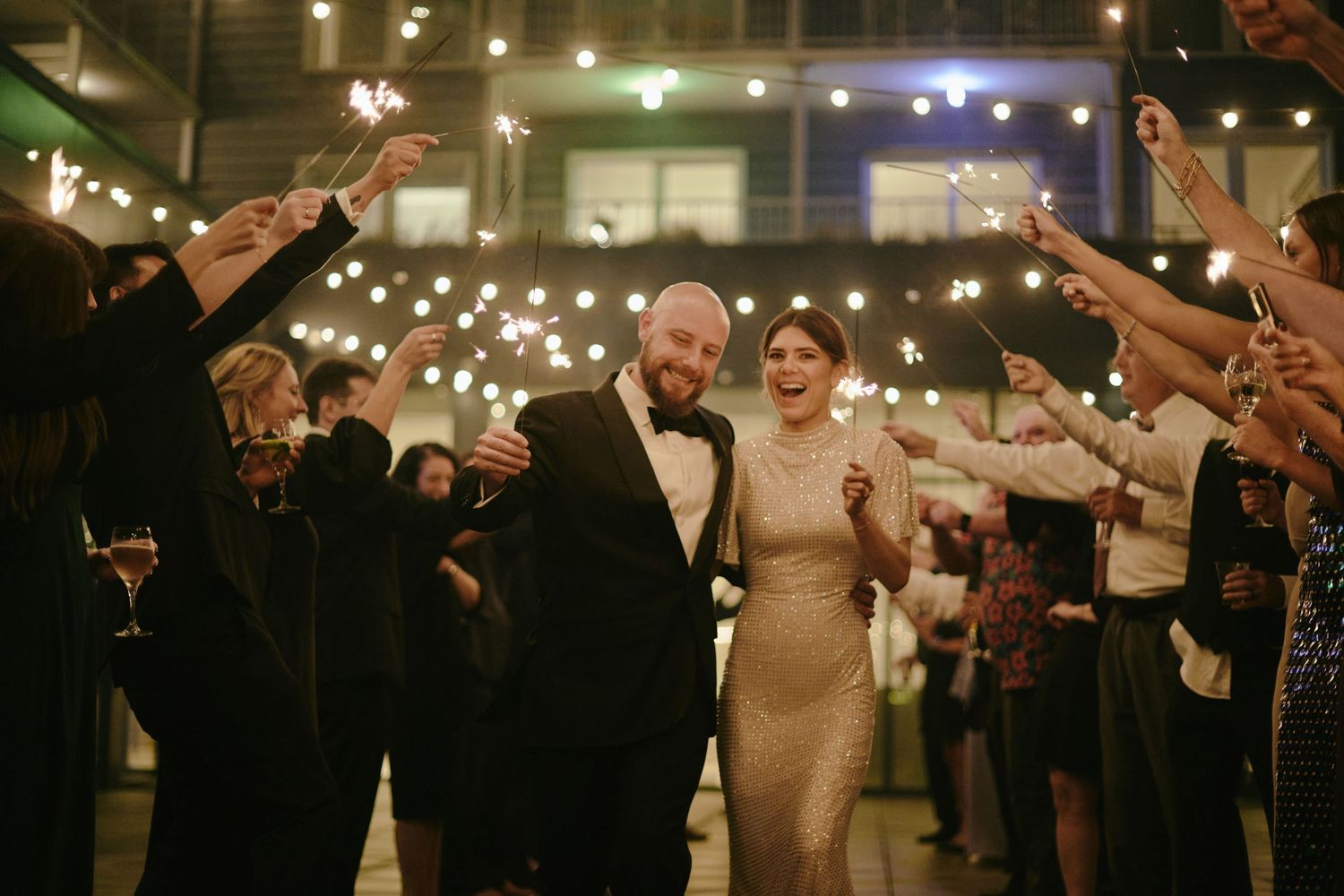 Bride and groom walk through sparkler tunnel surrounded by cheering wedding guests at elegant indoor reception.