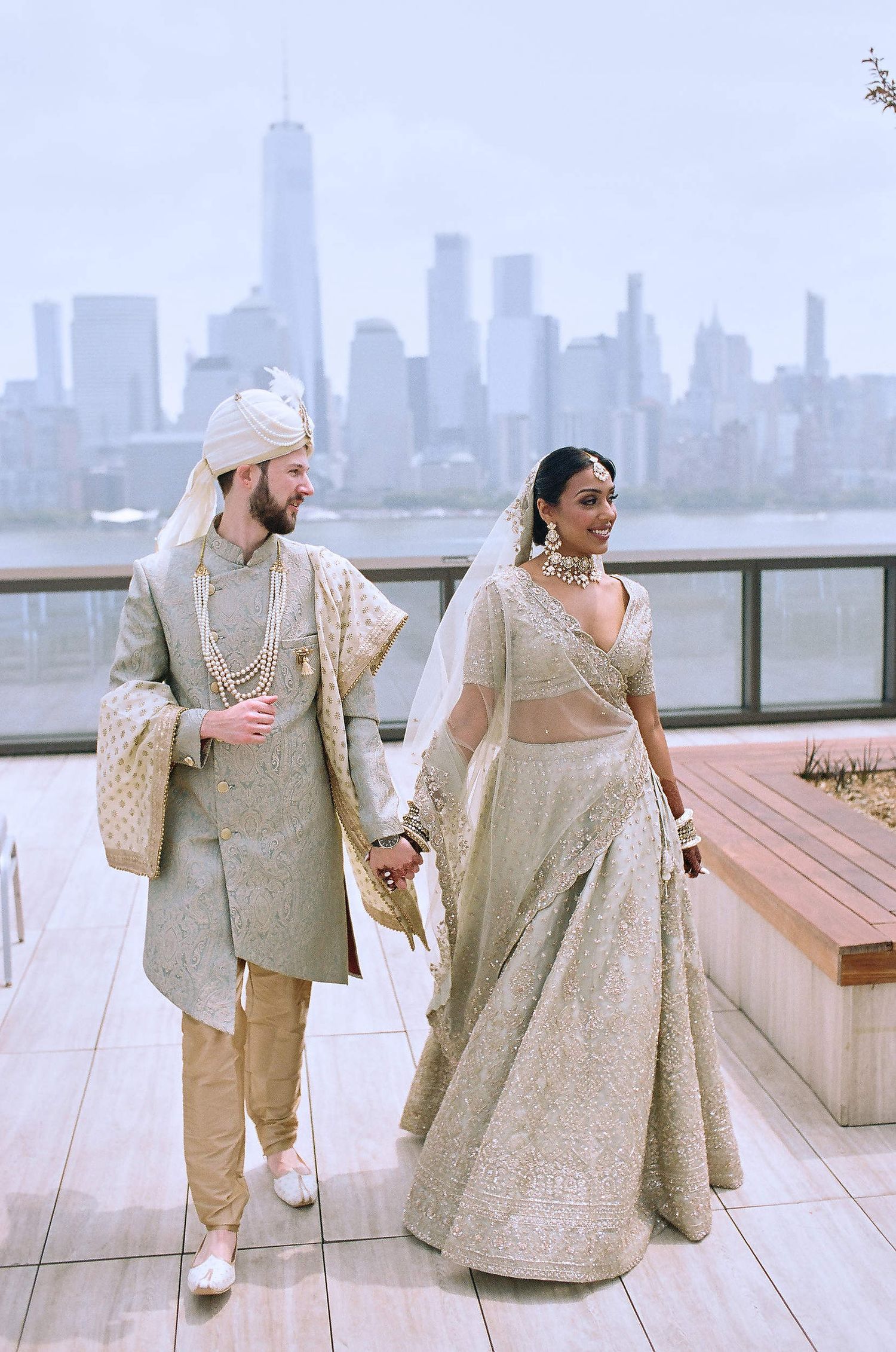 Traditional Indian wedding couple in elegant attire pose together on rooftop terrace with NYC skyline view in background.