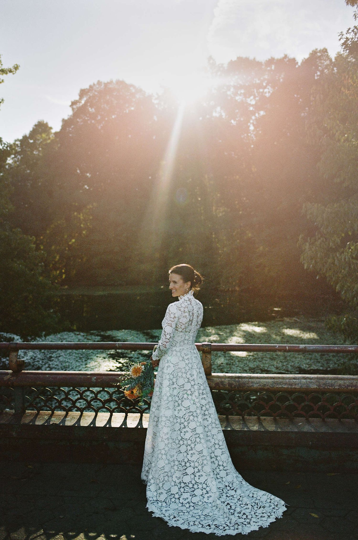 A person in an elegant white lace wedding dress stands on a bridge at sunset with sunbeams streaming through the trees.