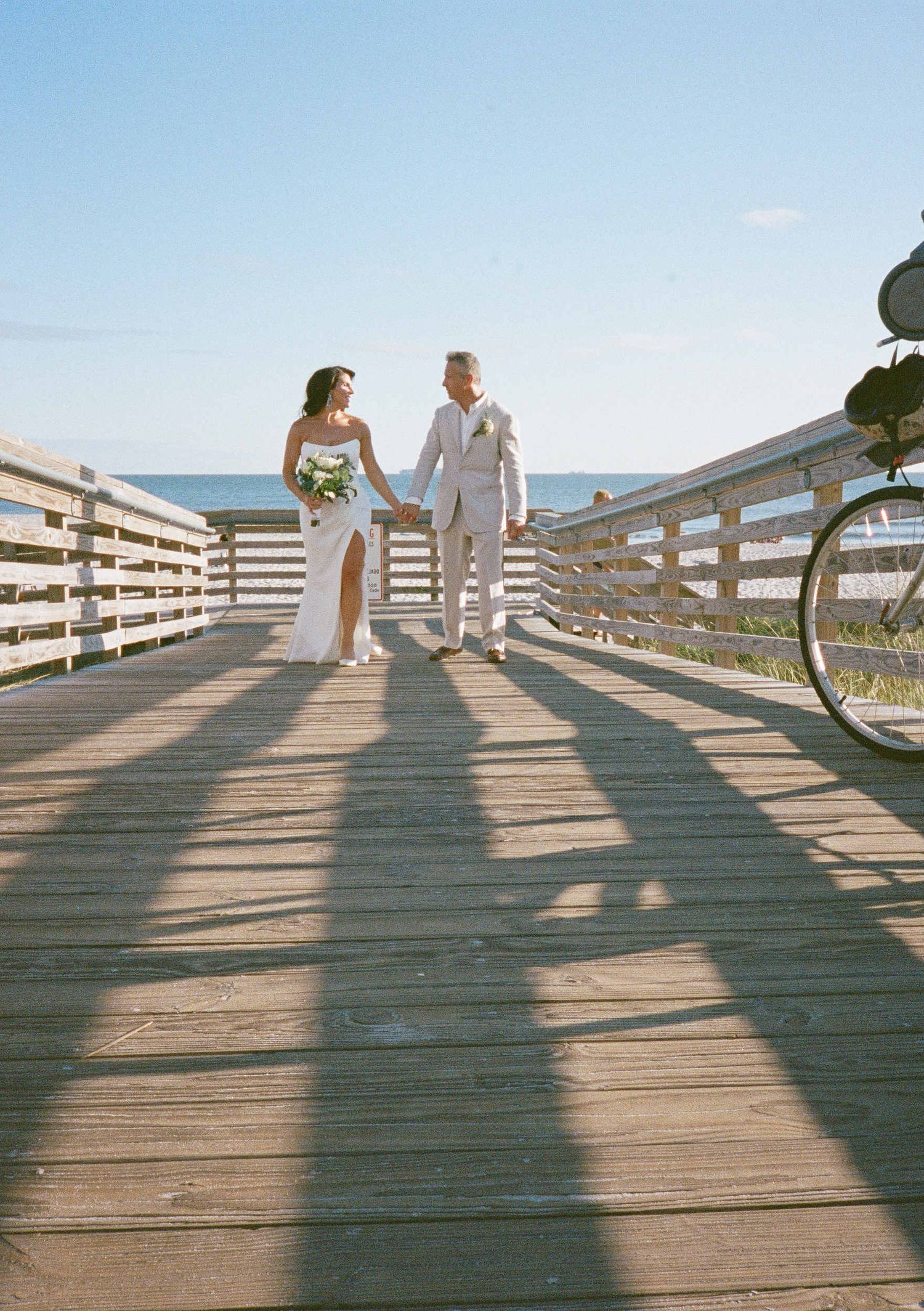 Couple holding hands while walking on a wooden boardwalk by the beach on their wedding day with long shadow cast on deck.