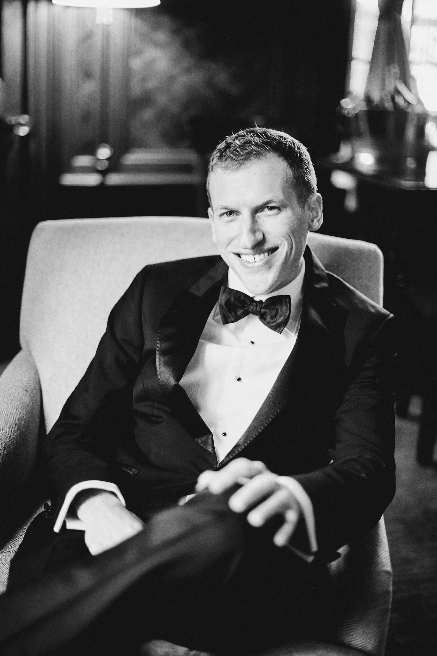 Black and white portrait of a smiling well-dressed man in a tuxedo and bow tie sitting in a chair.