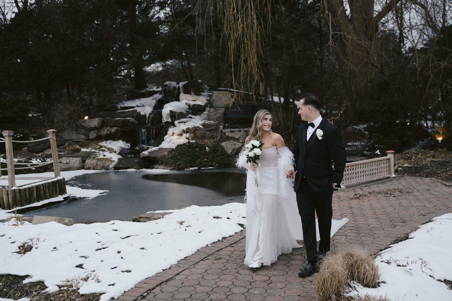 A bride and groom pose together on a snowy winter path with landscaped rocks and trees in the background.