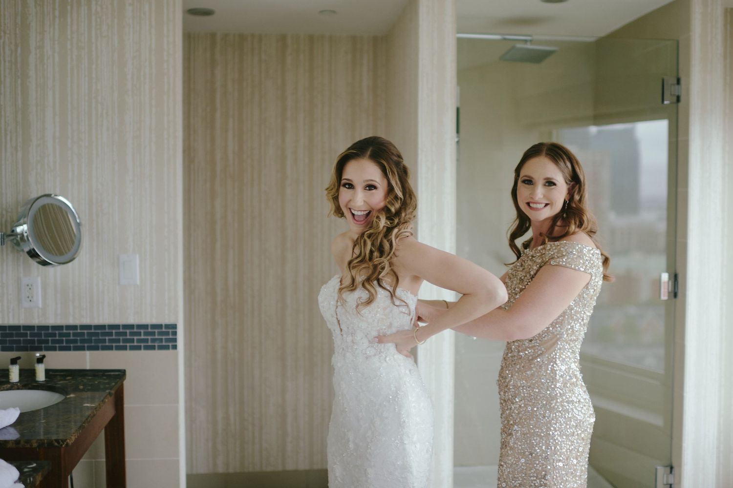 Two women in formal attire getting ready in an elegant hotel bathroom with beige walls.