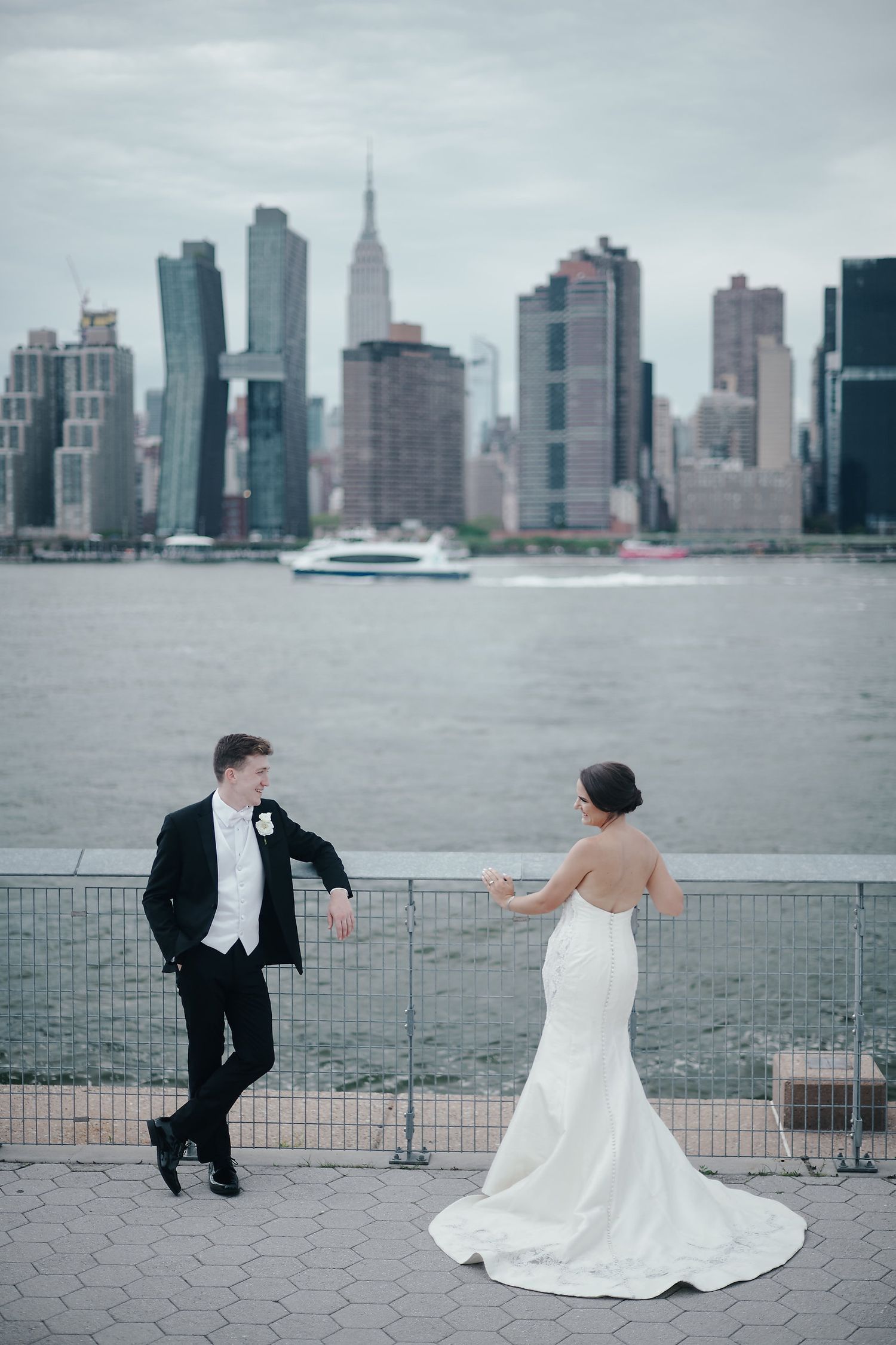 Couple in wedding attire dances playfully by waterfront with Manhattan skyline and ferry in background.