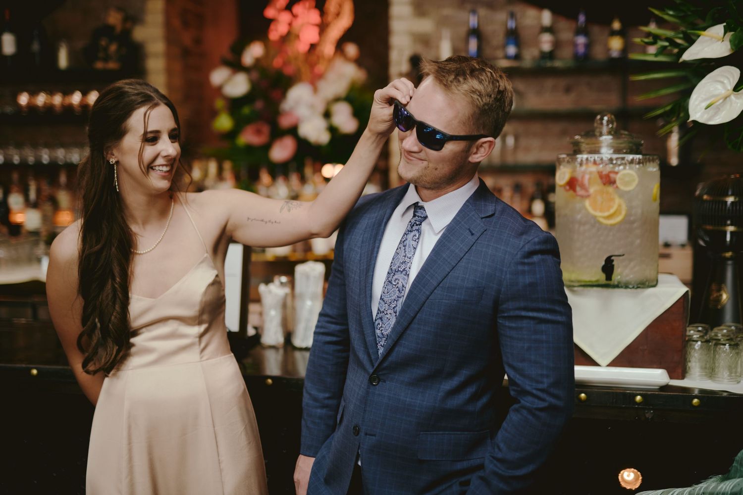 Two guests in formal attire share a playful moment by a bar decorated with pink flowers and a drink dispenser.