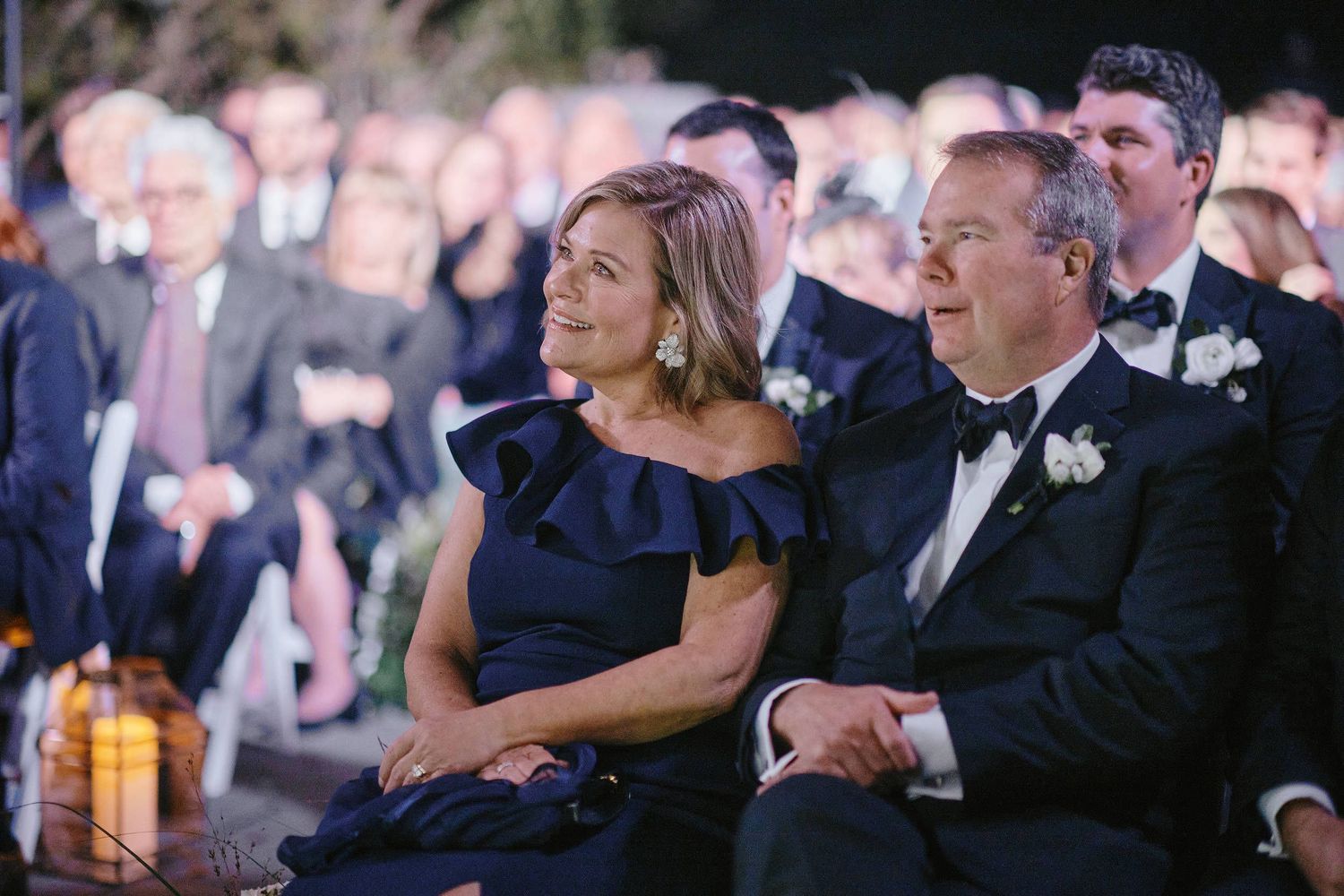 Well-dressed couple sits attentively in formal evening attire at an elegant event with a large crowd behind them.