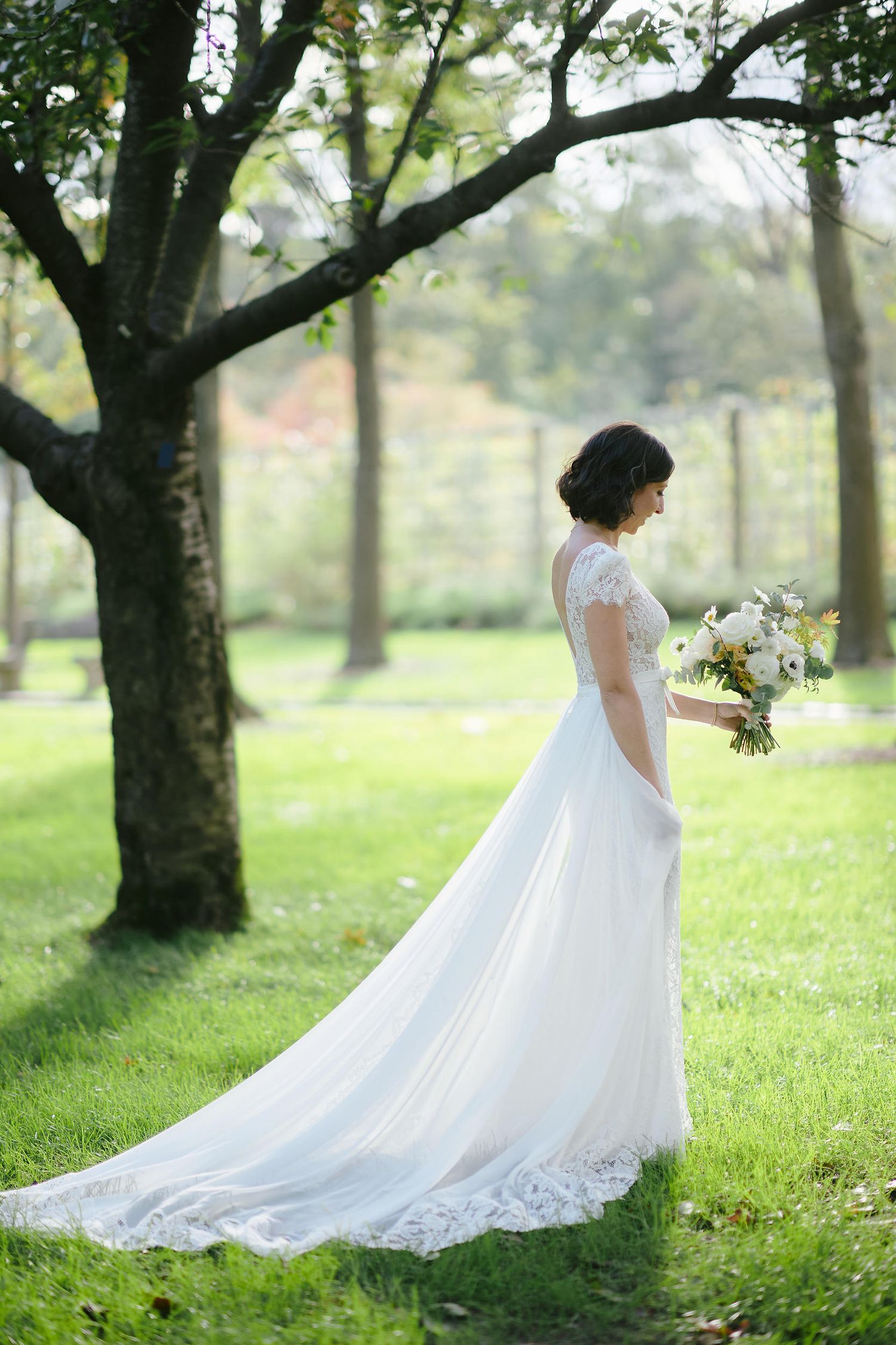 A bride in a white lace gown stands on lush grass holding a floral bouquet surrounded by trees in an outdoor setting.