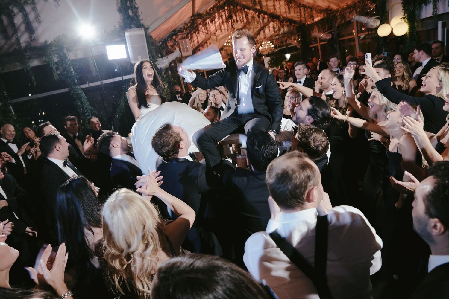 Wedding guests lift and carry a couple above the crowd during a lively reception celebration at an indoor venue.