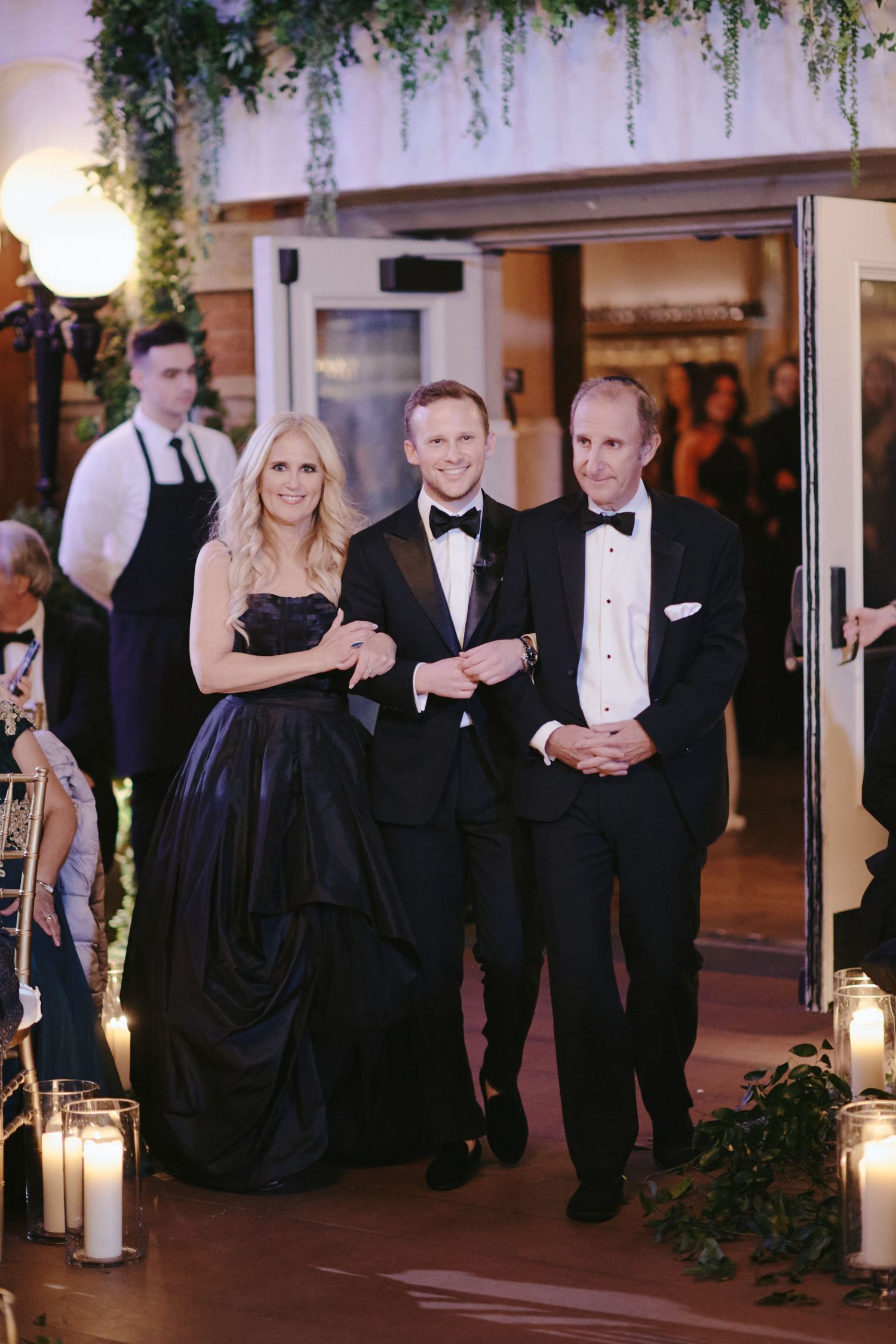 Elegantly dressed guests in formal black tie attire enter a candlelit venue decorated with hanging greenery for an upscale event.