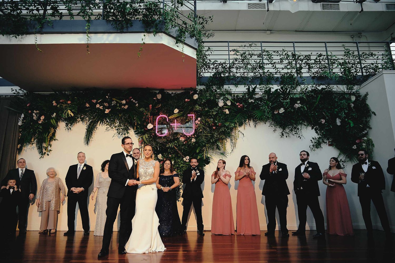 Wedding party in formal attire stands against greenery-decorated wall during reception at indoor venue.