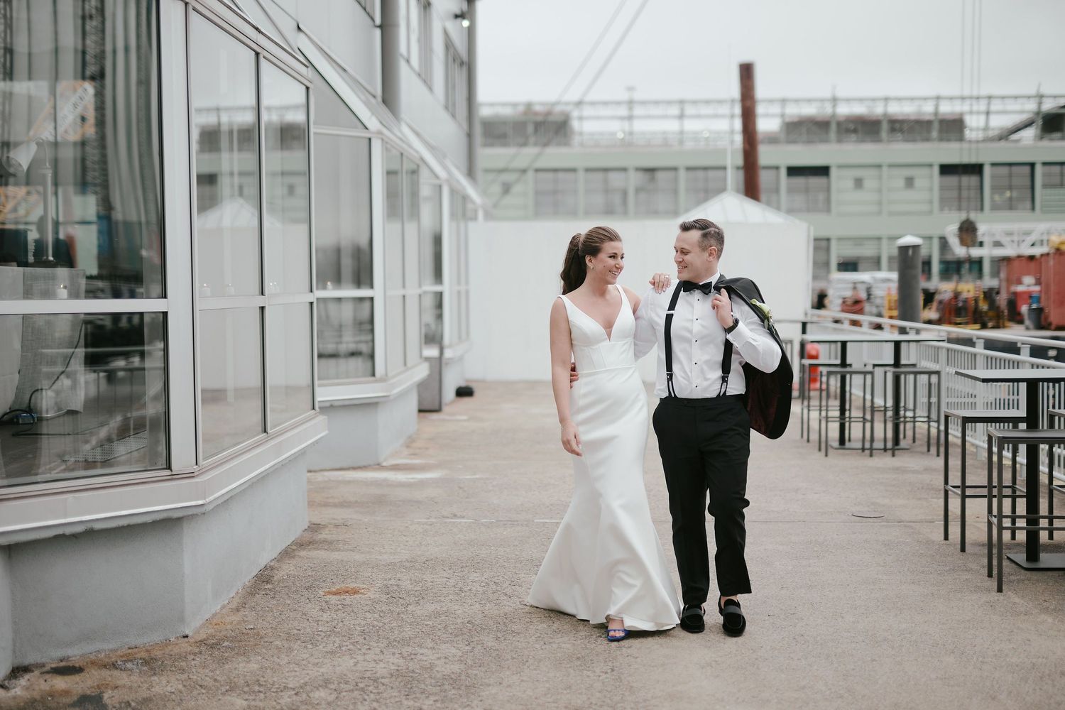 A couple walking together outside an industrial building on their wedding day, with her in white and him in a dark suit.