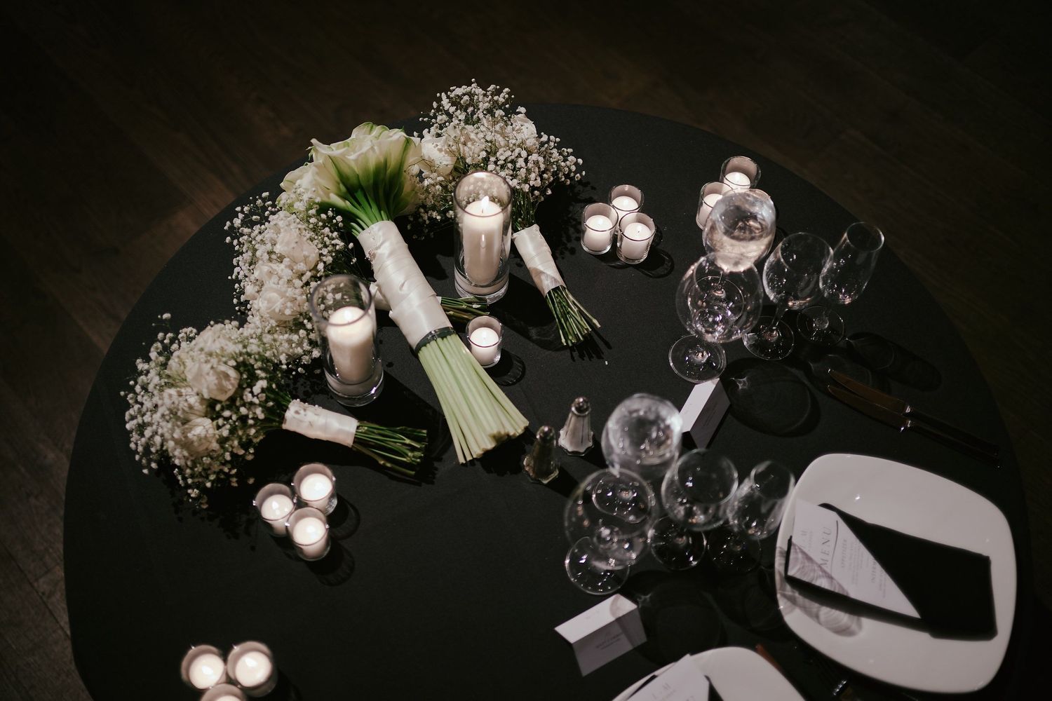 Wedding bouquet with white roses and baby's breath alongside glasses and table settings on dark surface.