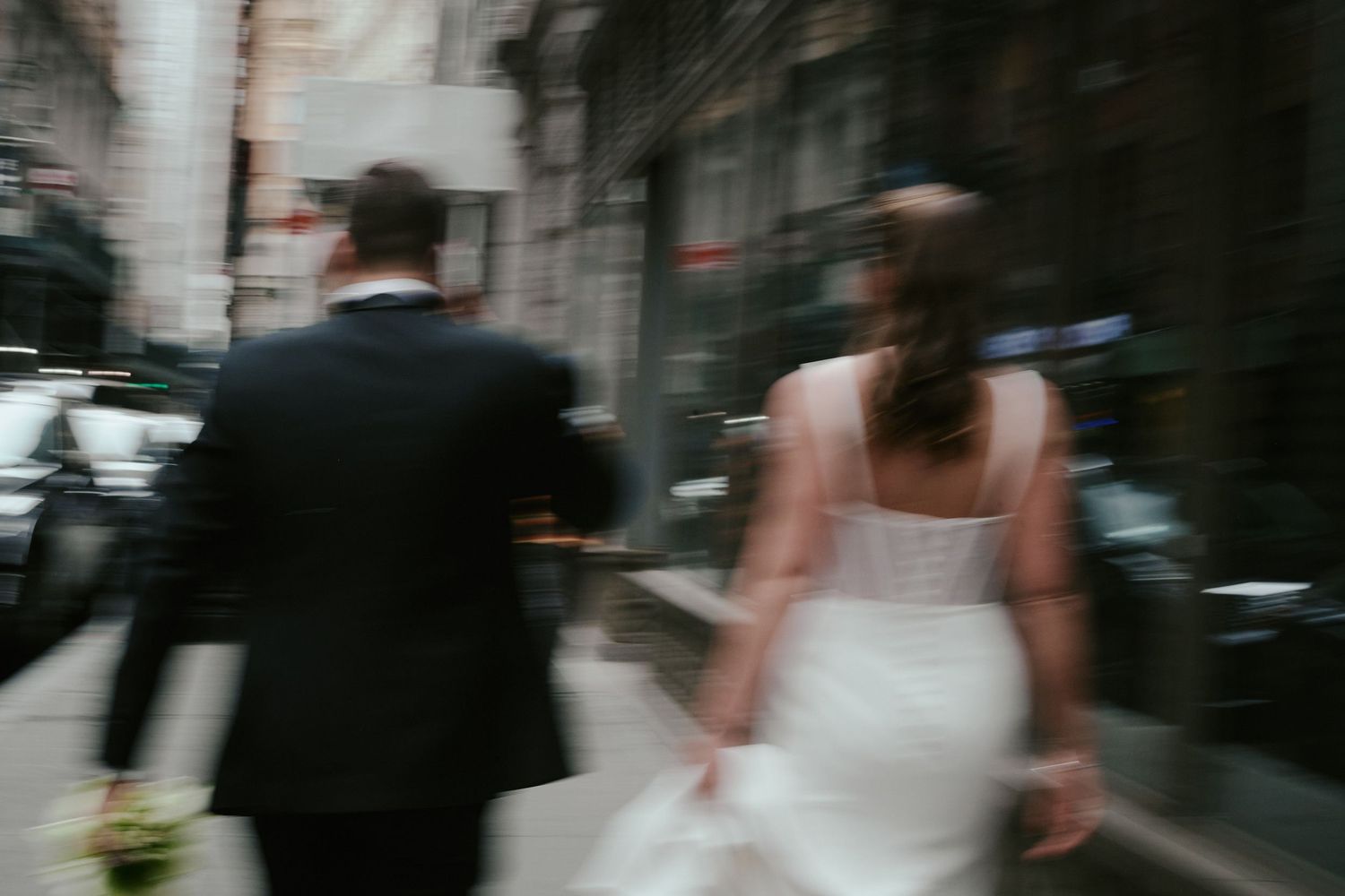 Blurred motion shot of a bride in white dress and groom in black suit walking down a city street.