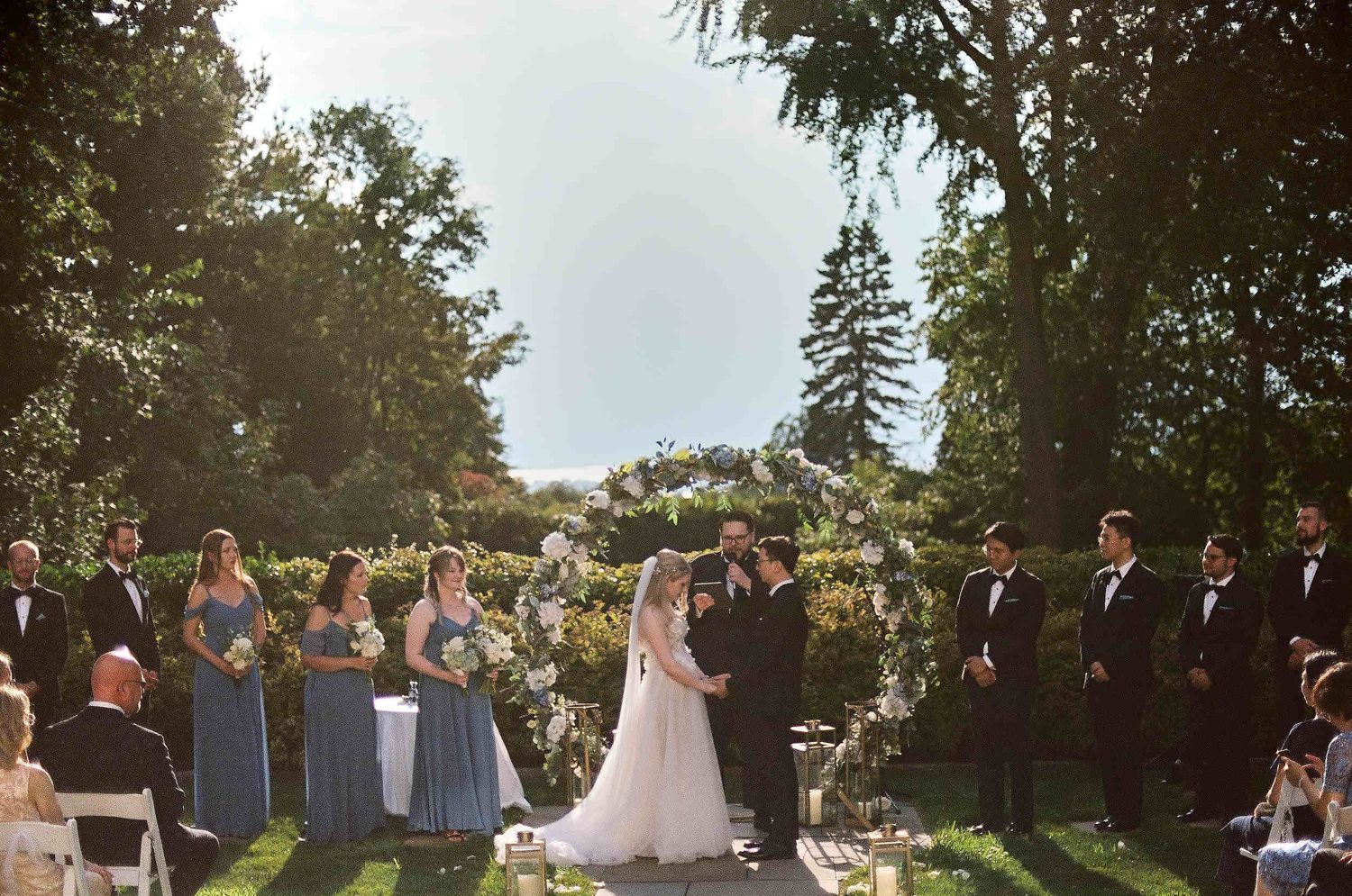 Wedding ceremony taking place in a garden setting with bridesmaids in blue dresses and groomsmen in dark suits.