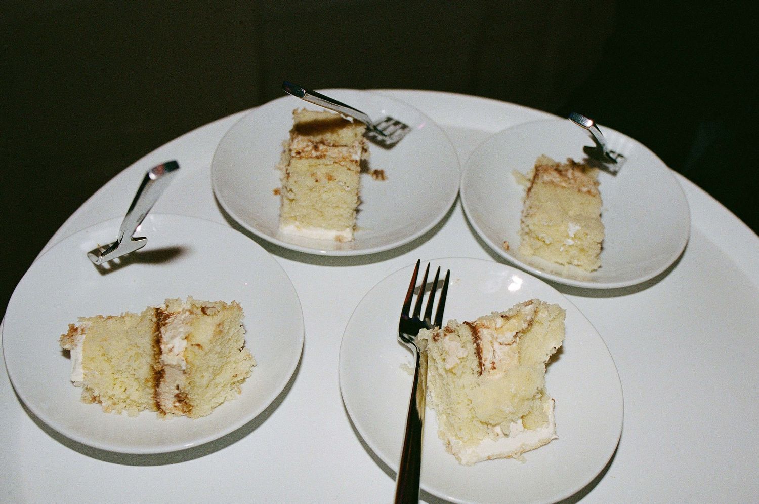 Four plates of vanilla layer cake slices with white frosting arranged on a white surface with a fork.