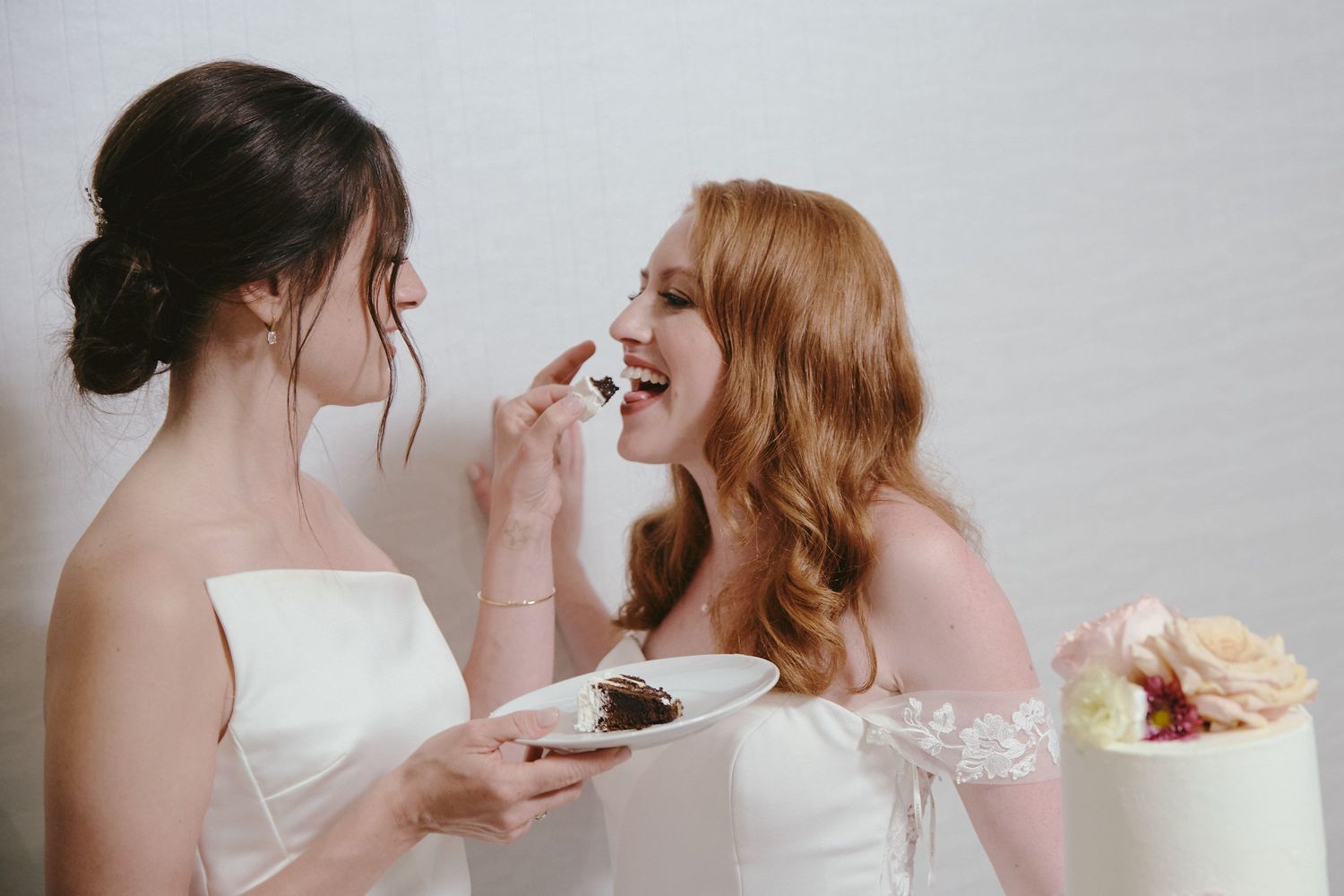 Two brides in white dresses playfully feed each other cake during their wedding celebration.