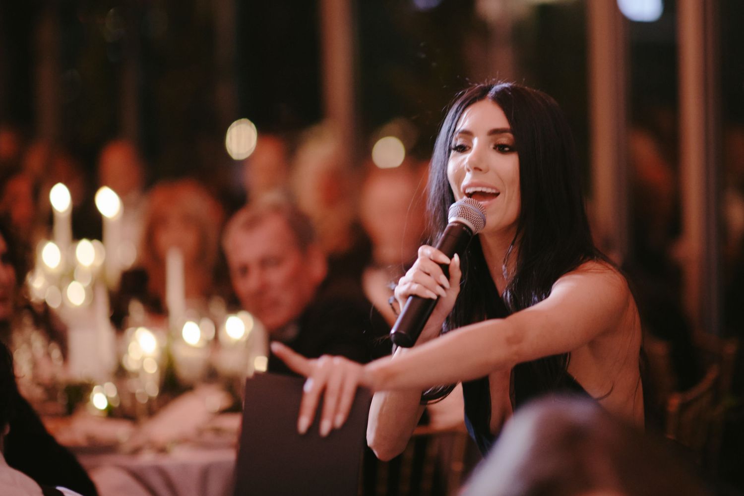 A wedding reception at night with lit candles on tables while someone is speaking into a microphone.