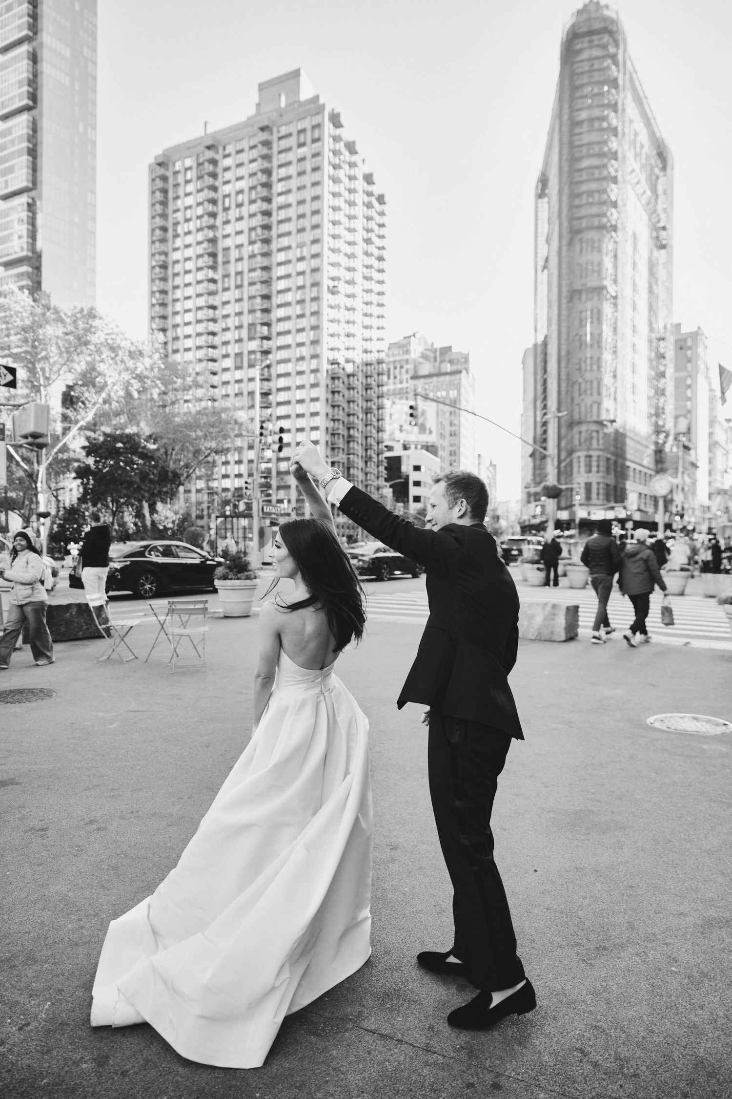 A couple in wedding attire dancing and twirling on a New York City street with the Flatiron building in the background.
