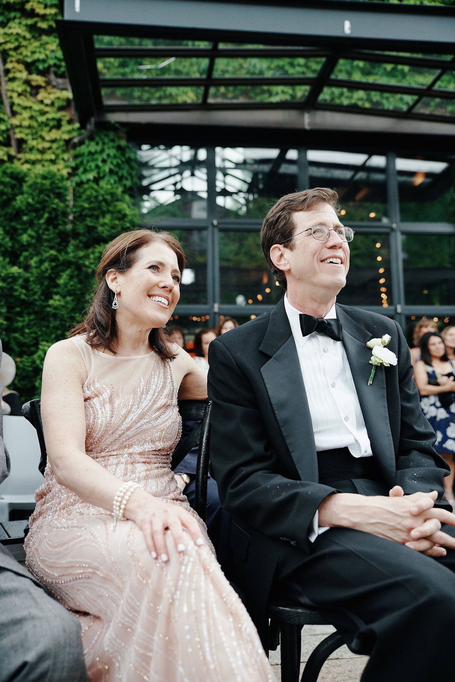 Couple seated at outdoor wedding venue wearing formal attire with greenhouse and greenery backdrop.