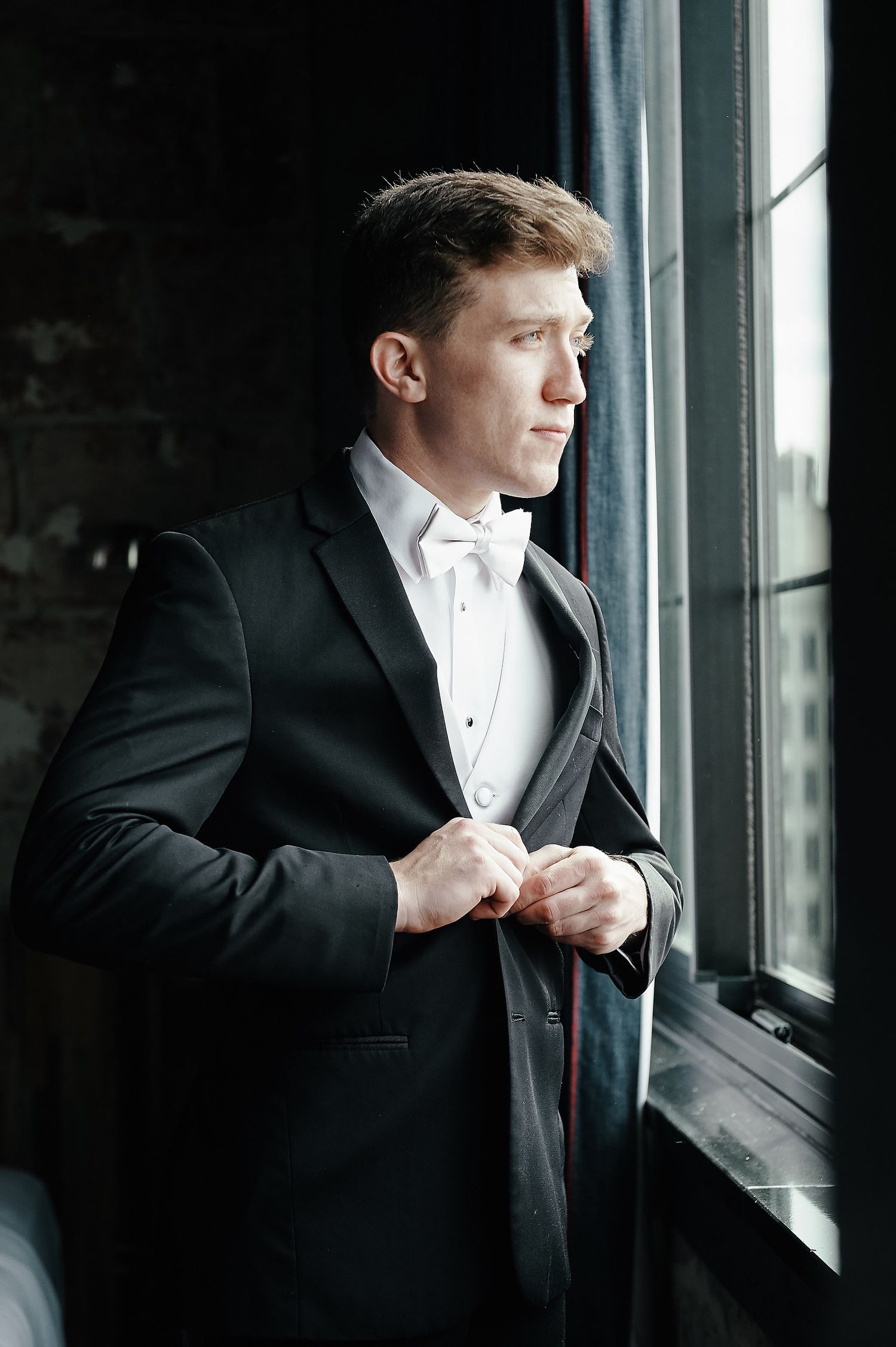 A person in a formal black tuxedo and white tie stands thoughtfully by a window in natural light.