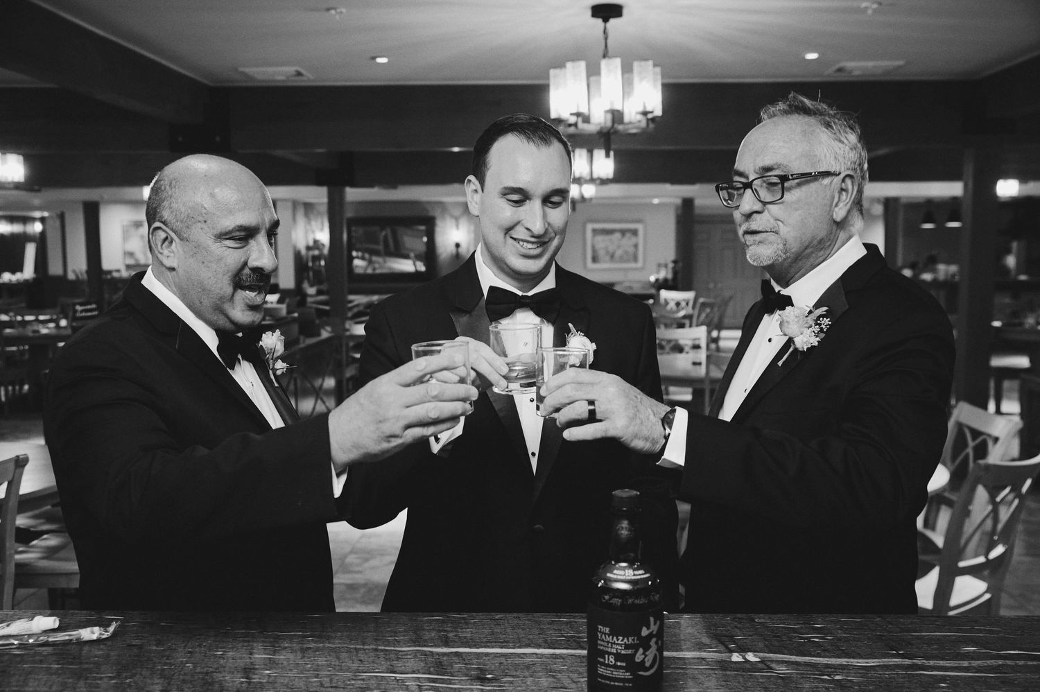 Three men in formal attire toast with drinks at an elegant bar counter in a black and white photograph.