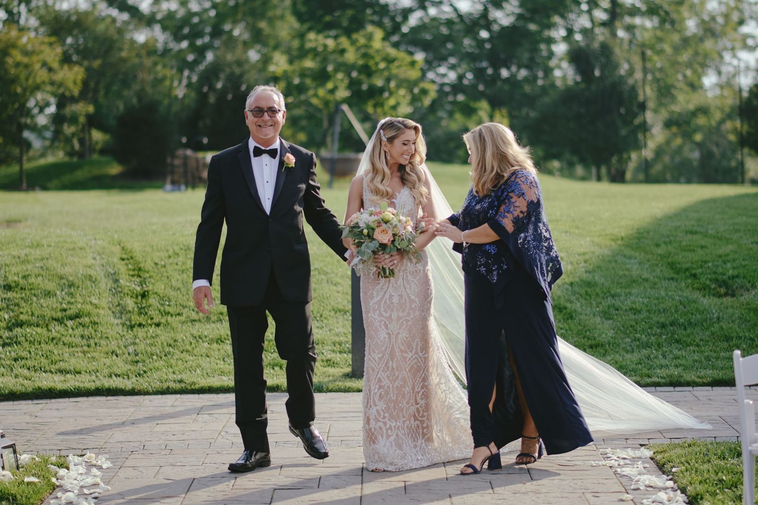 Three people in formal attire stand on a stone path outdoors during what appears to be a wedding celebration.