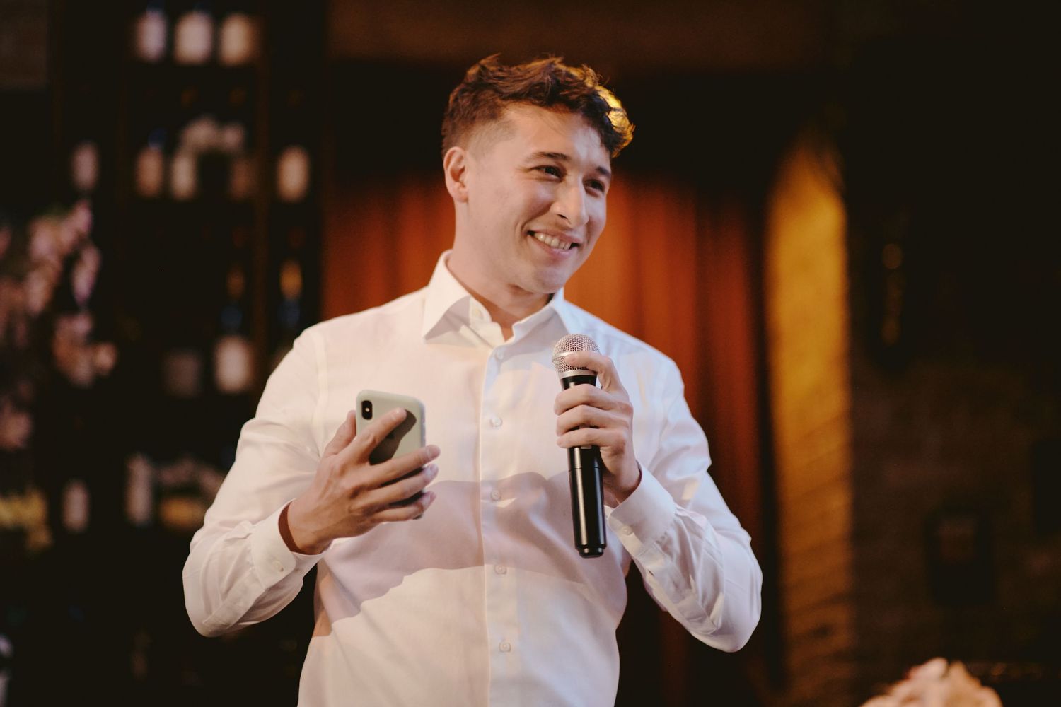Young speaker in white shirt holds microphone while giving speech at indoor evening event.