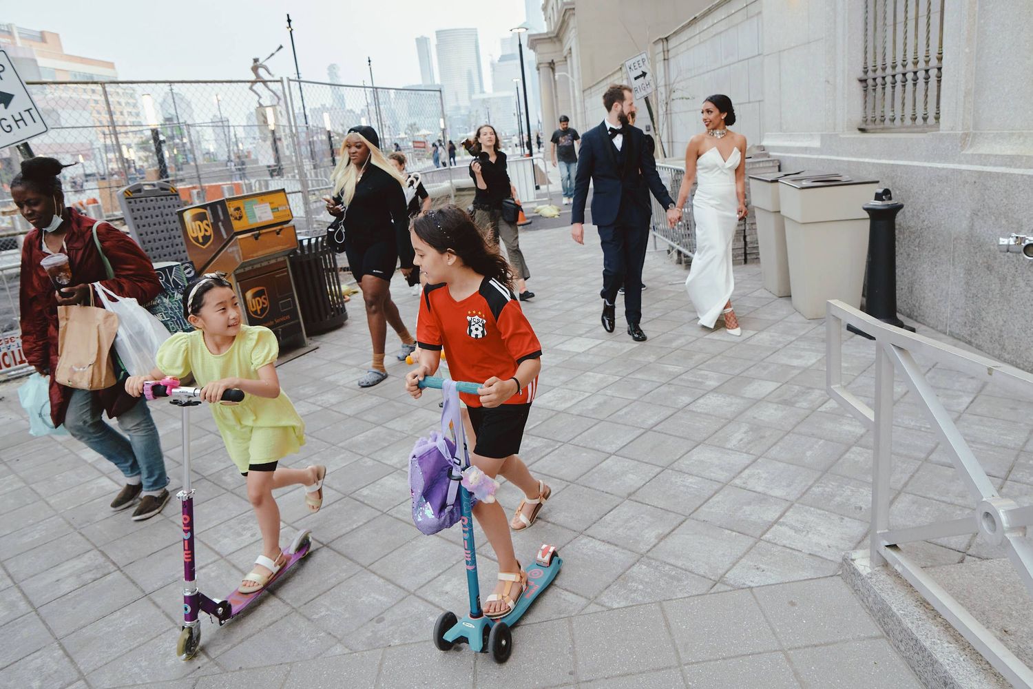 Children riding scooters on city sidewalk while wedding party walks behind them on urban street.