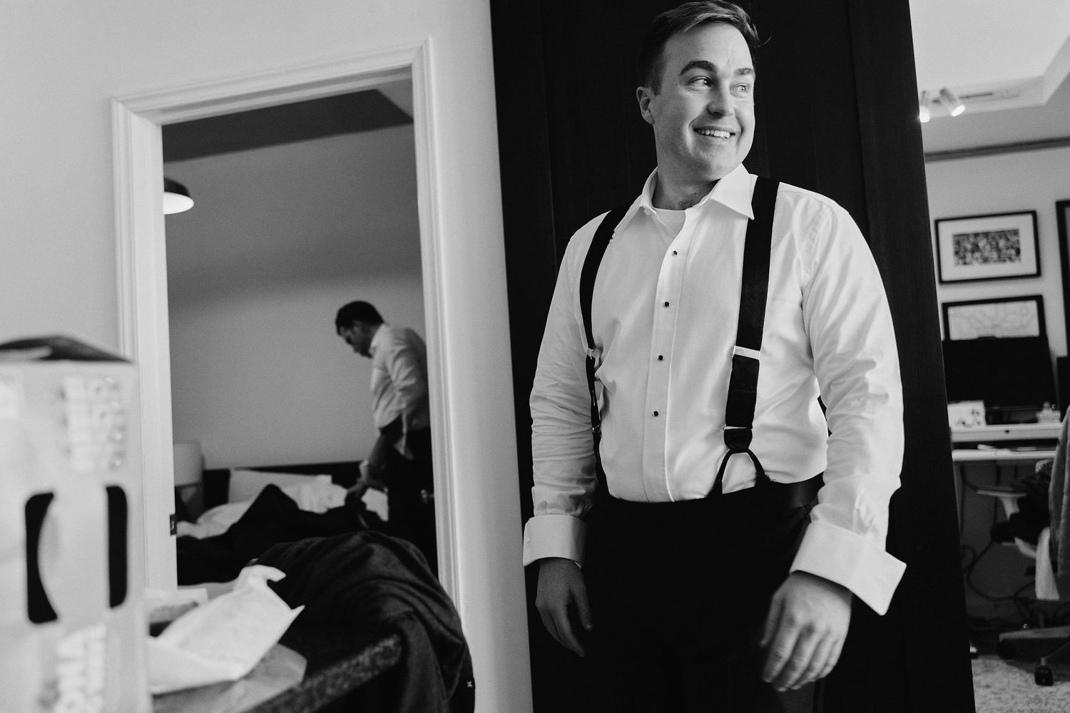 Black and white photo of a smiling person in formal attire with suspenders standing in a doorway.
