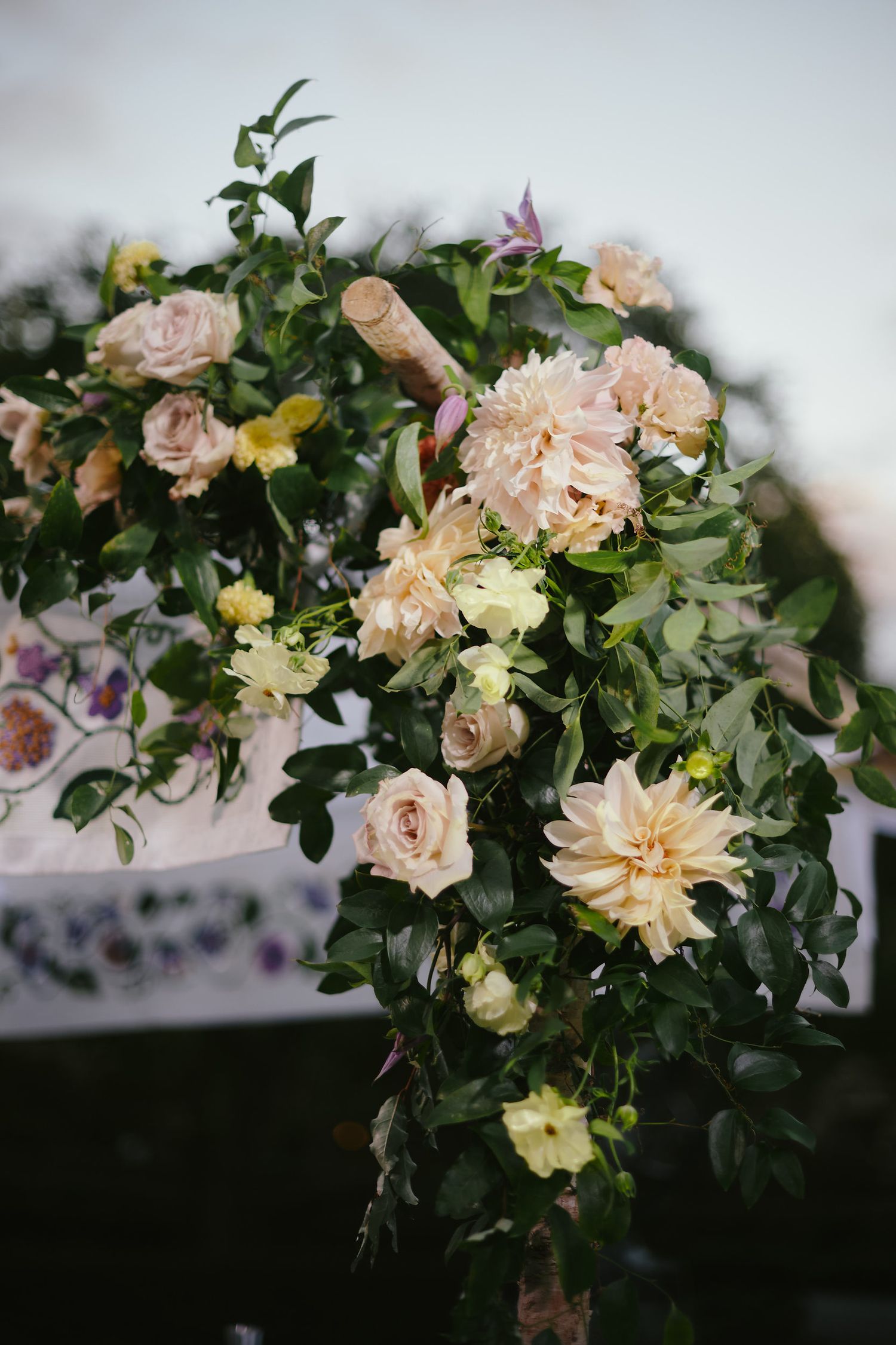 A lush floral arrangement featuring peach roses, pale dahlias and yellow blooms mixed with green foliage in soft focus.