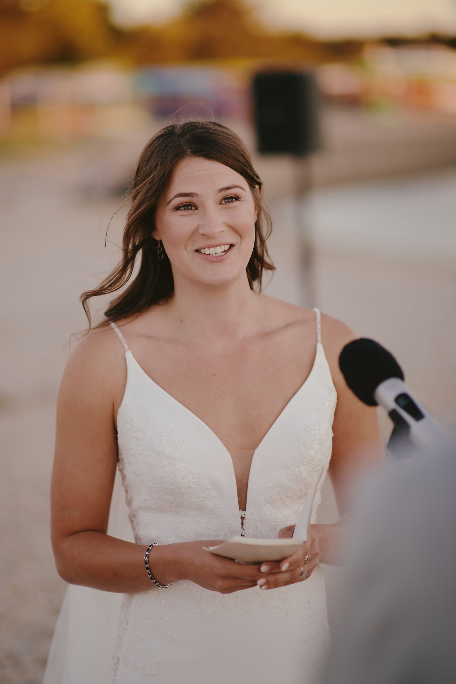 Someone in a white v-neck wedding dress smiles while holding a microphone during an outdoor ceremony.