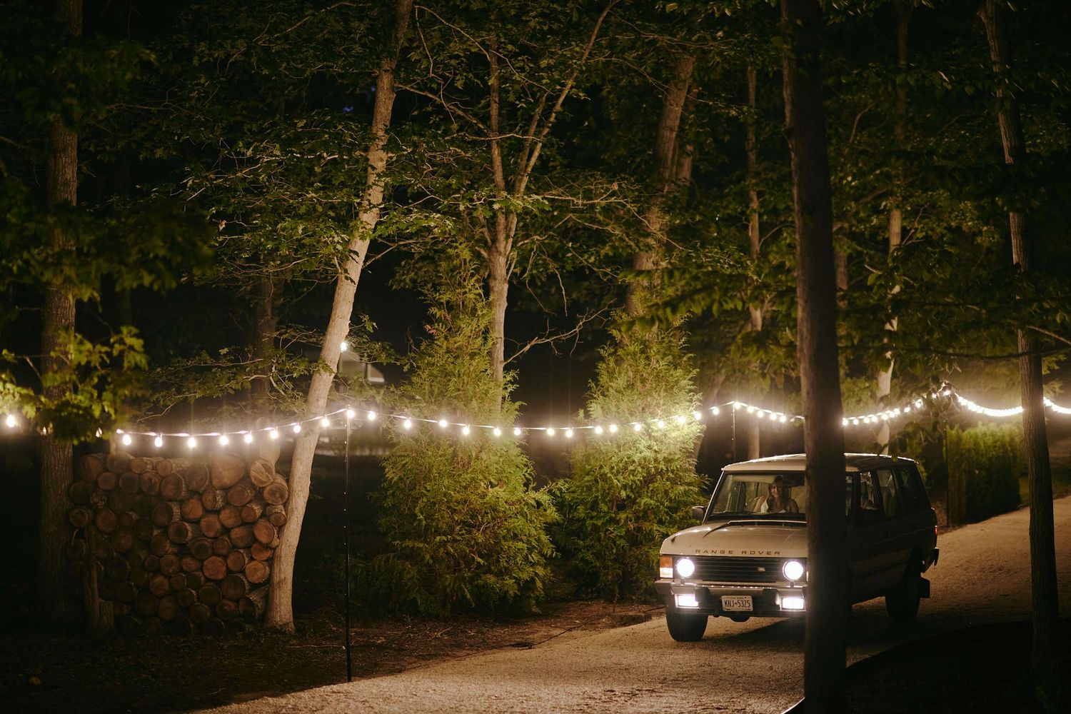 String lights illuminate a dark wooded area with a parked vehicle beneath the glowing canopy.