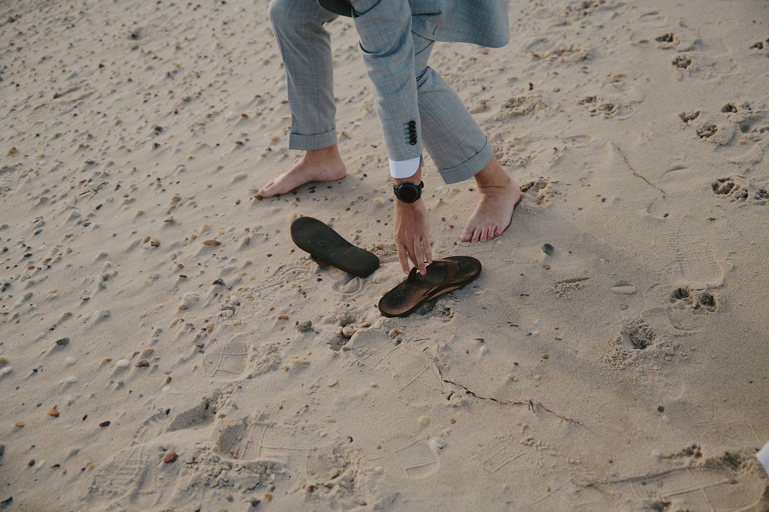 Bare feet stand next to black flip flops on sandy beach, wearing rolled up blue jeans.