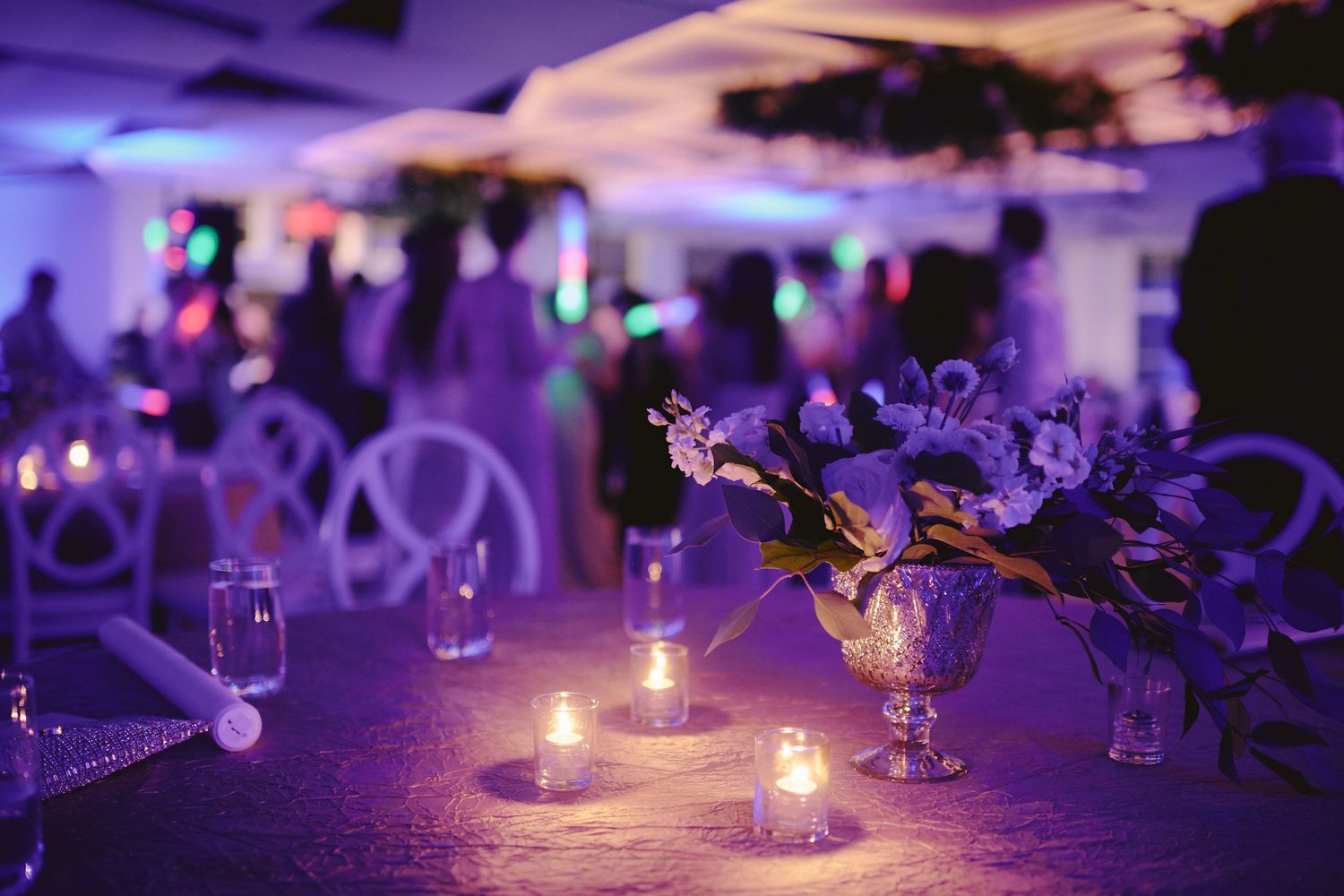 An elegant wedding reception table with purple lighting, glowing candles and floral centerpieces in a luxurious venue.