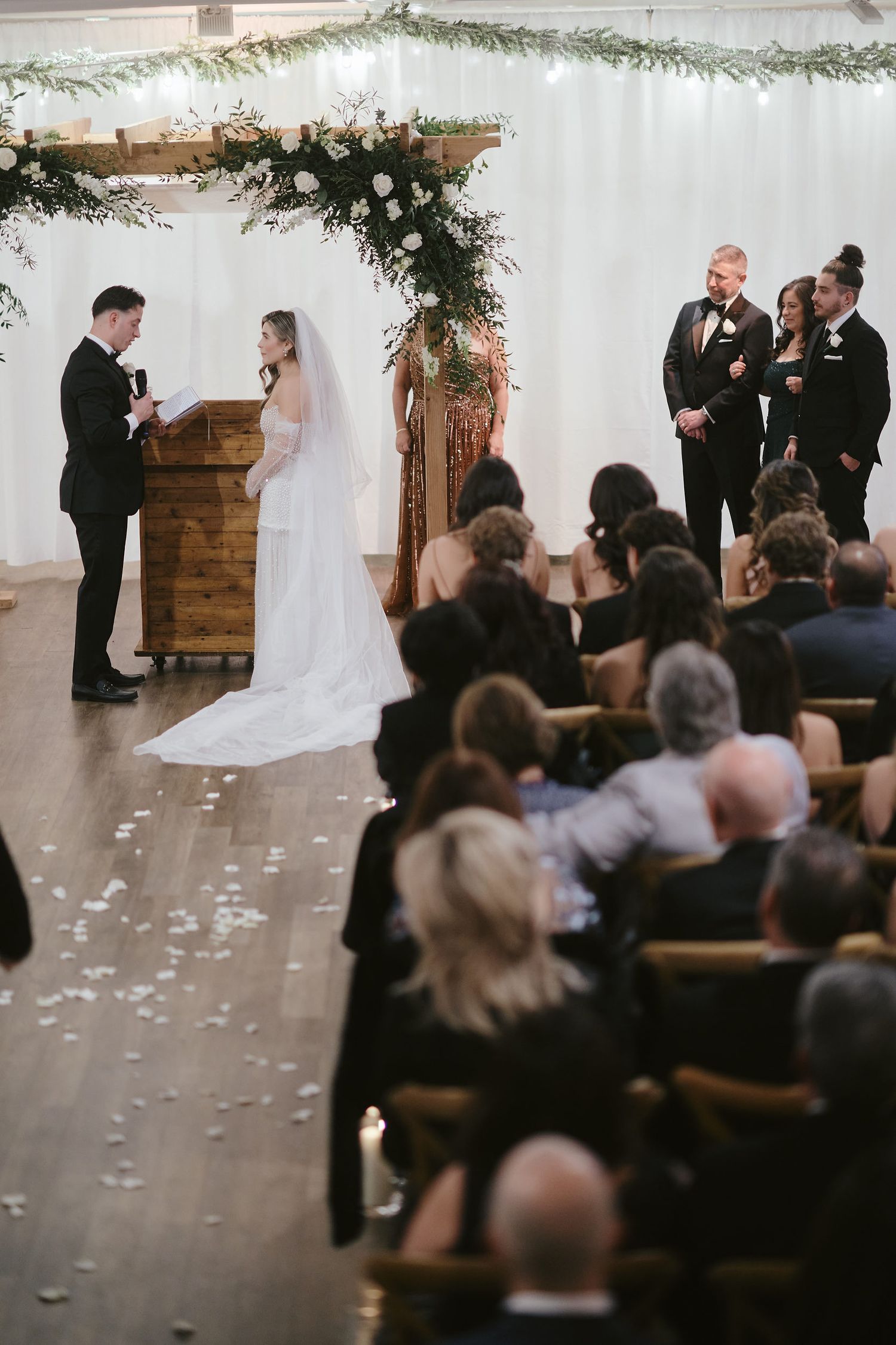 Wedding ceremony taking place indoors with floral arch and guests seated in wooden chairs.