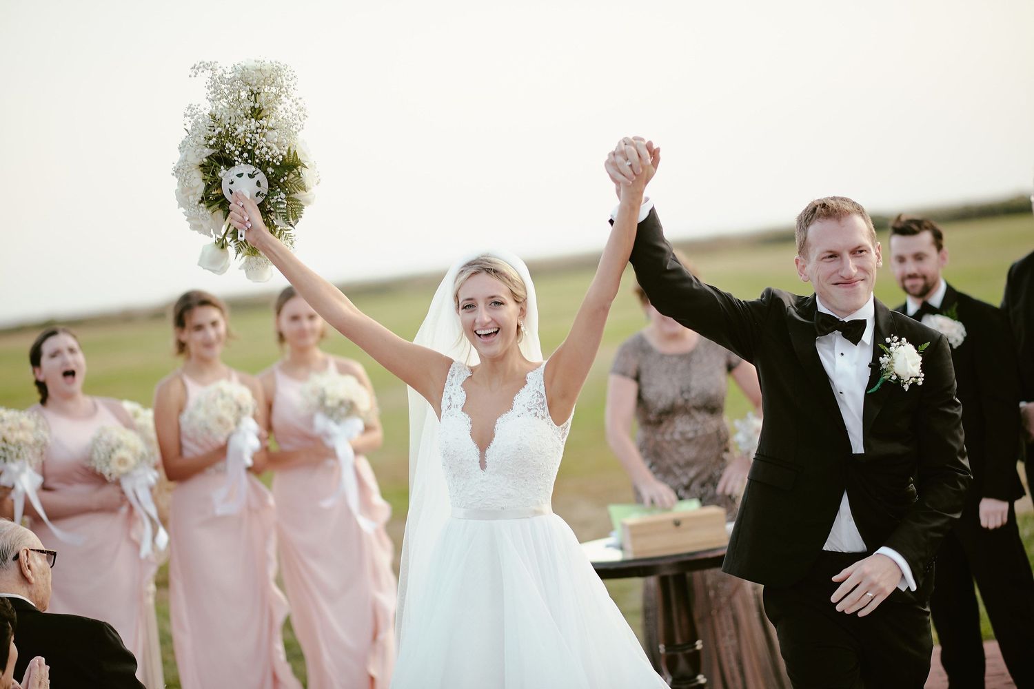 Joyful newlyweds raise their hands in celebration while bridesmaids in pink dresses applaud at an outdoor wedding ceremony.