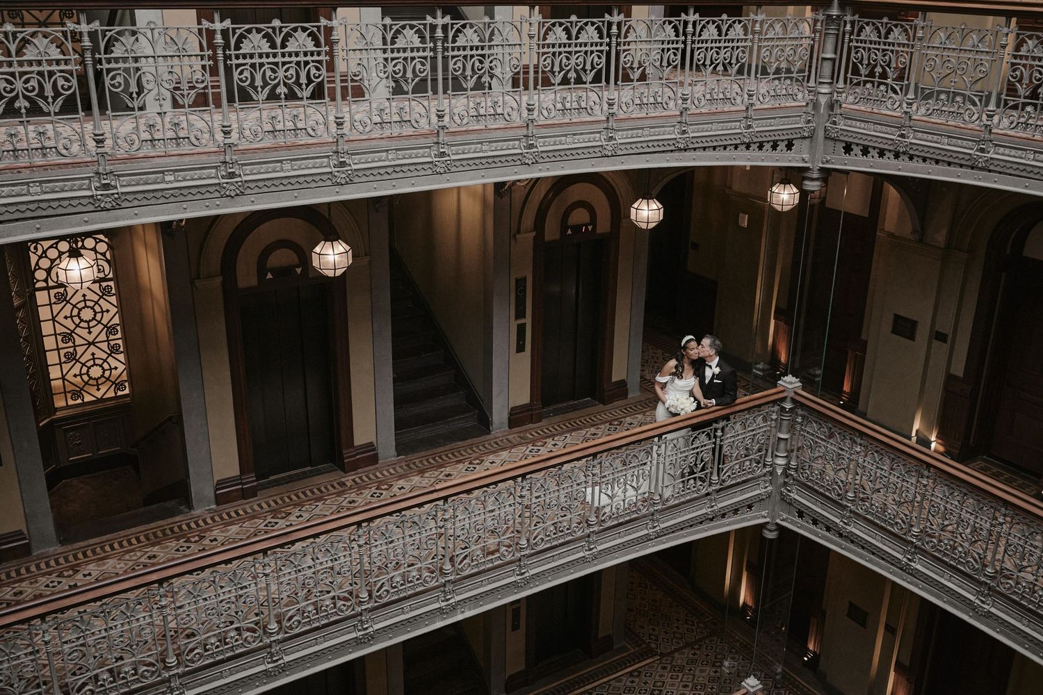 Elegant wedding couple on ornate balcony of historic multi-story venue with decorative iron railings and chandeliers.