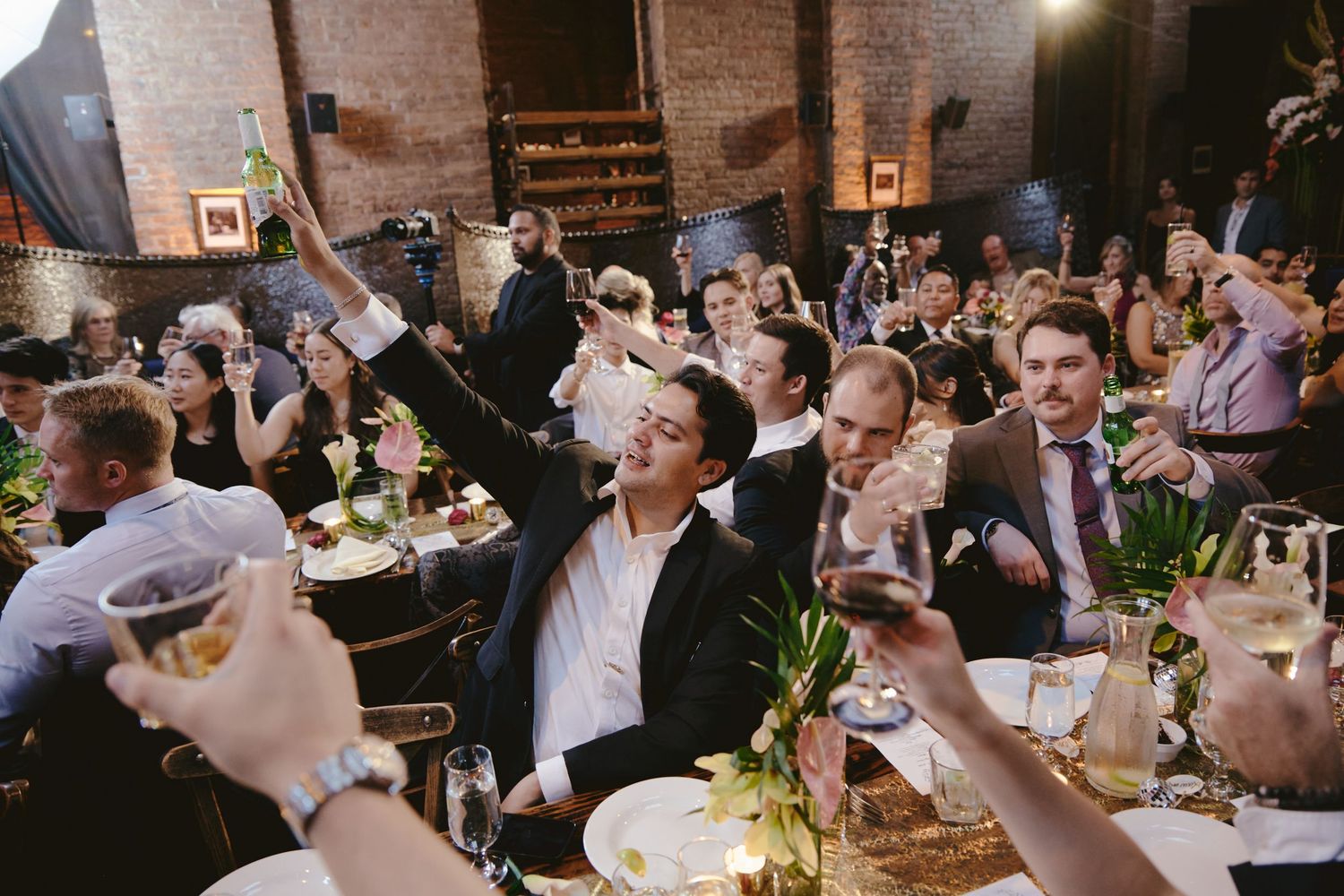 Wedding guests celebrate joyfully at a rustic brick-walled reception venue with raised glasses and lively energy.