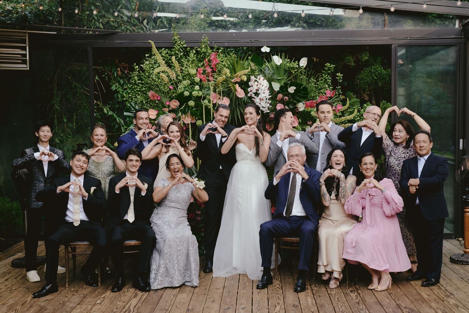 A festive wedding party poses together on a wooden deck while making heart shapes with their hands.