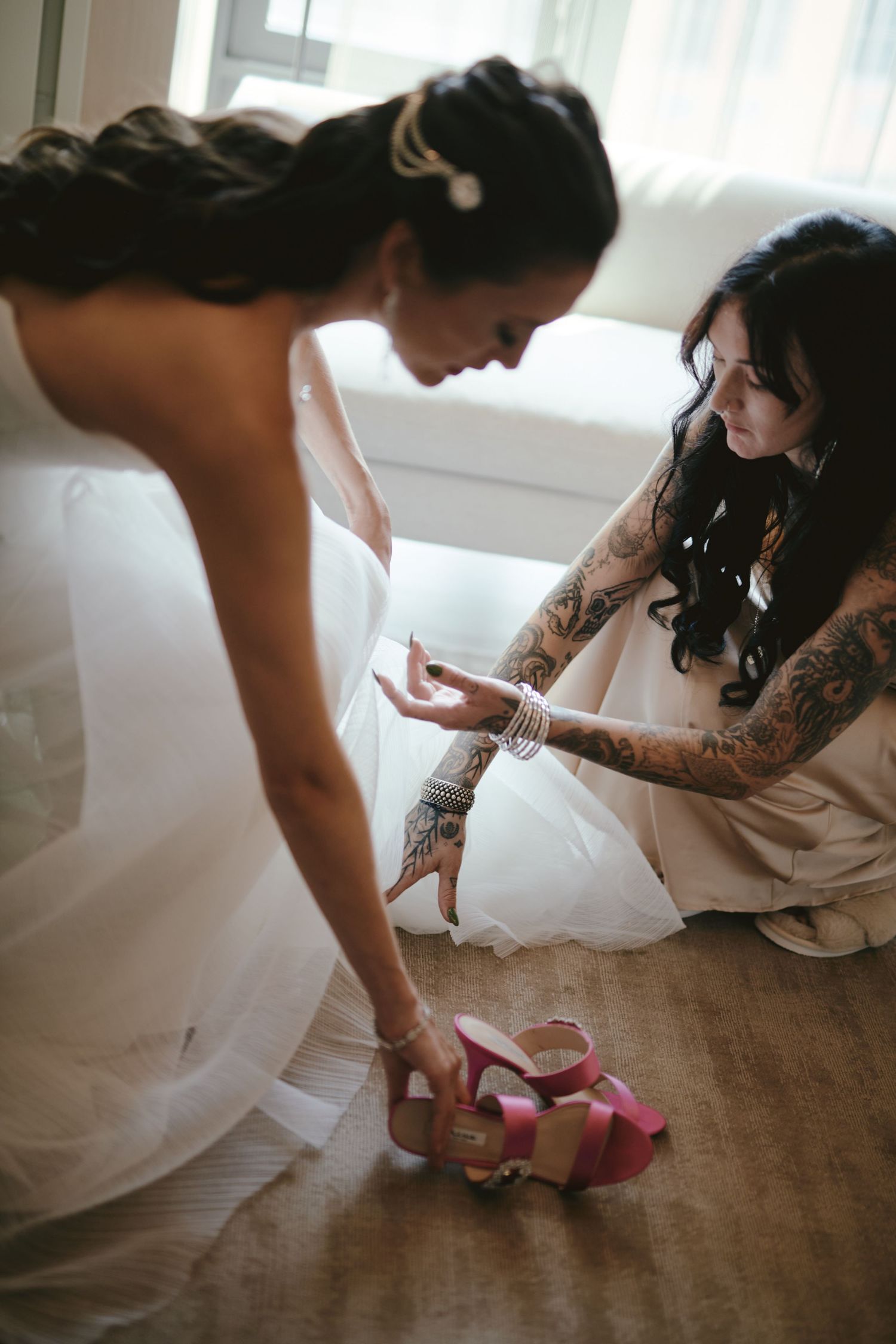 A person in a white wedding dress tries on pink platform heels while seated on the floor with a companion.